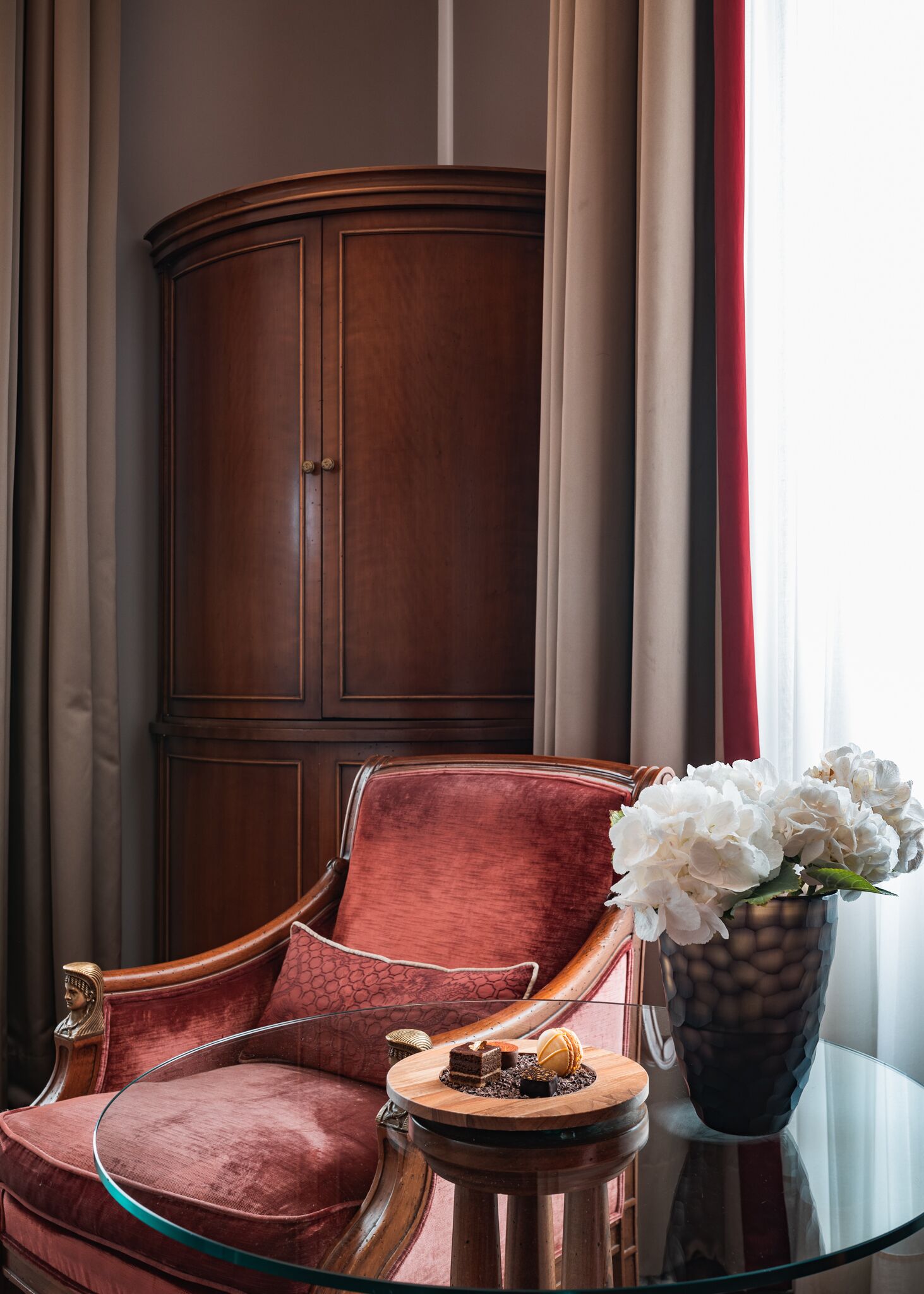 A room with a wooden cabinet, a red upholstered chair, a glass table with a vase of flowers, and a glass coffee table with desserts.