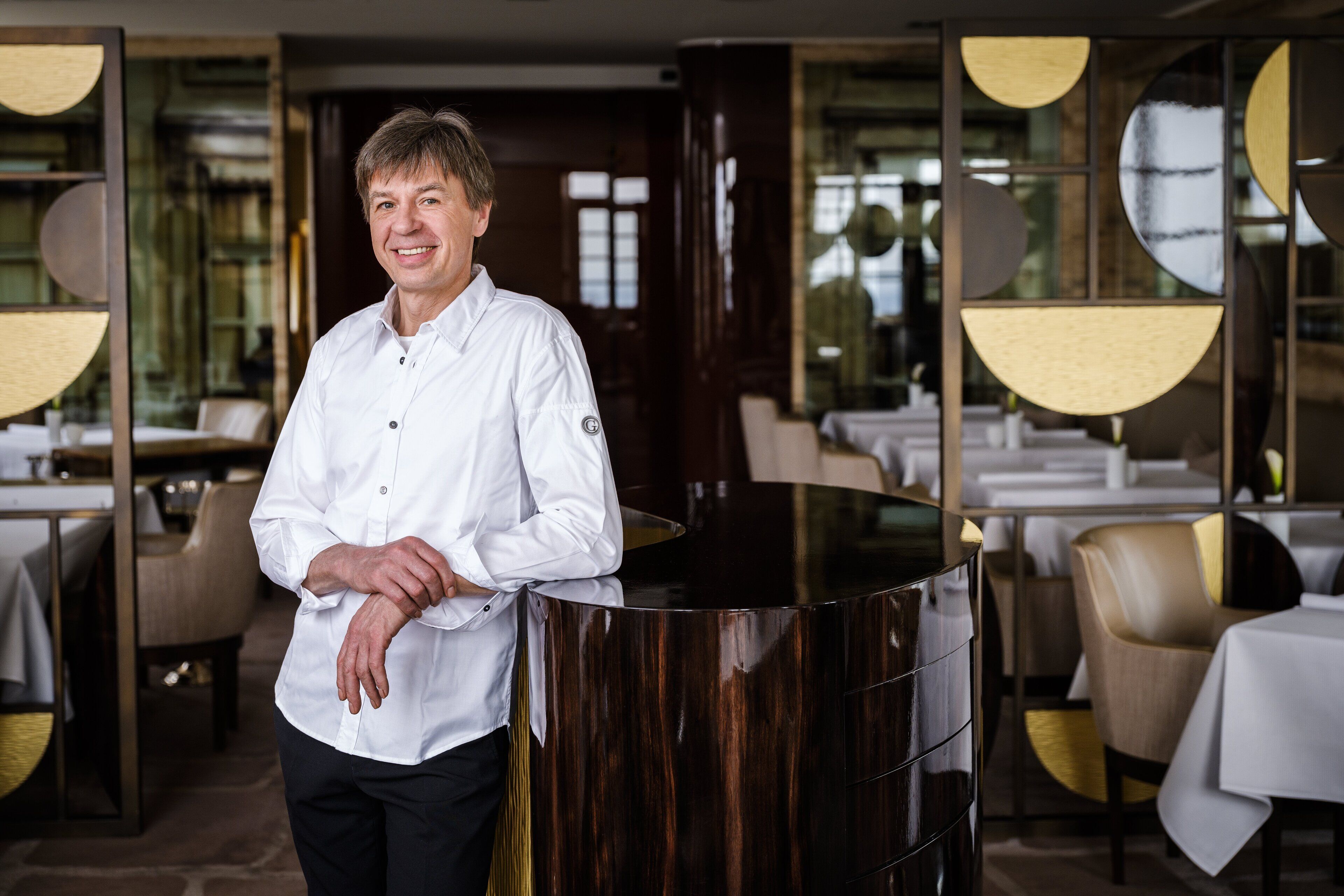 A man in a white shirt standing by a polished black counter in a modern dining area with tables and chairs.