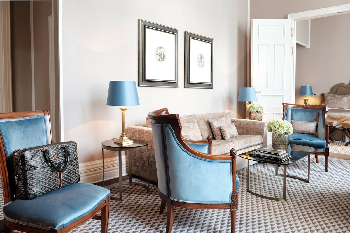 Una sala de estar elegantemente decorada con un sillón azul, un sofá y una mesa de centro. Hay dos cuadros enmarcados en la pared, una lámpara sobre una mesita auxiliar y una puerta al fondo.