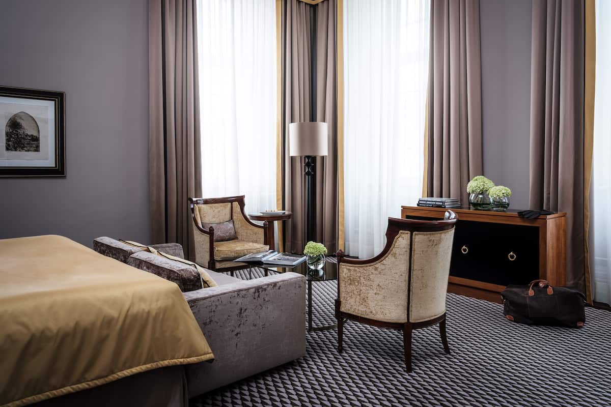 Habitación de hotel con cama, sofá, sillón, escritorio con lámpara y una consola decorada con objetos ornamentales.