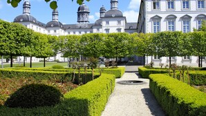 Un jardin à la française, caractérisé par une disposition symétrique, qui mène à un imposant bâtiment doté de plusieurs dômes et d’un style architectural classique.