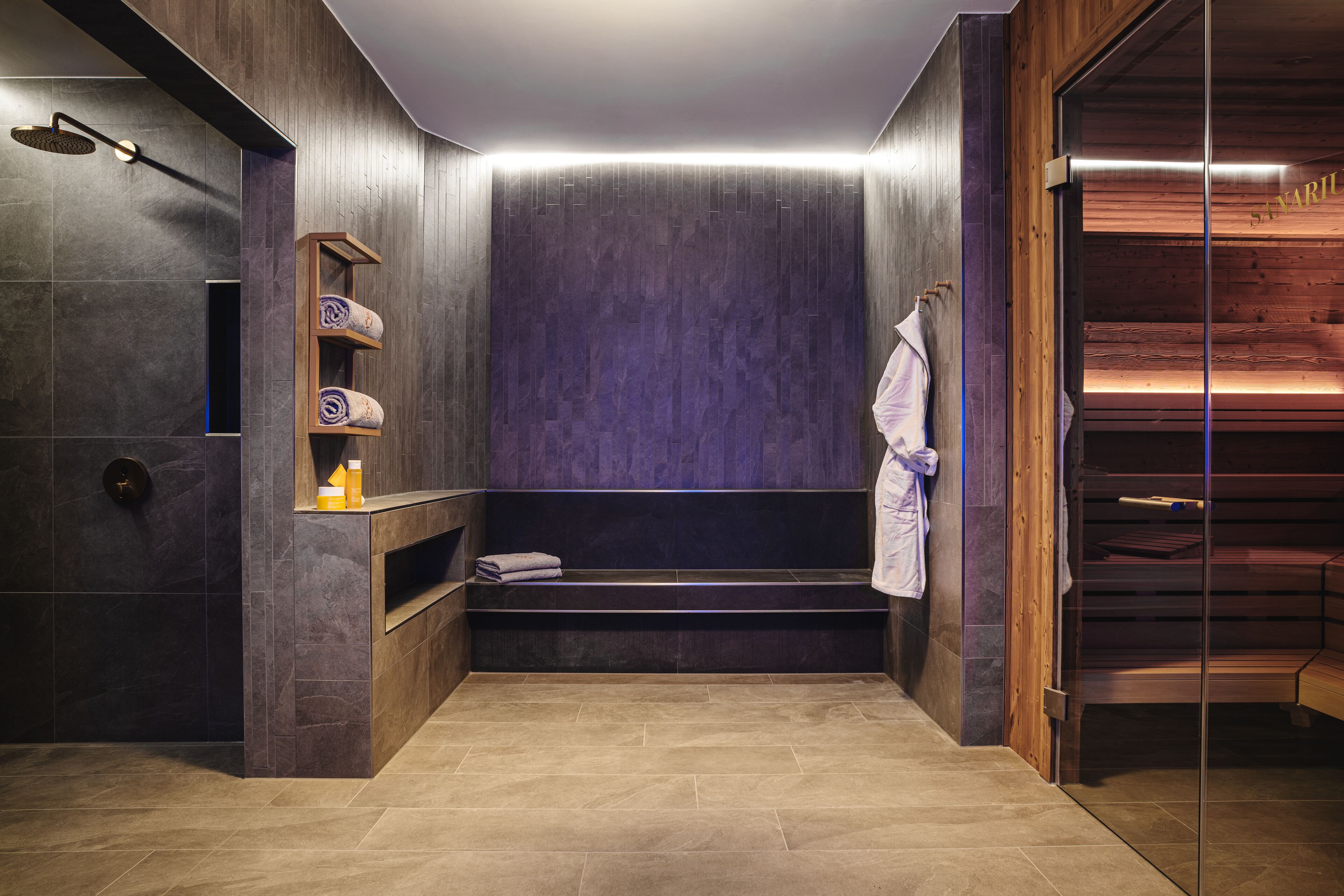 Un baño moderno con ducha integrada, repisas de madera con toallas y un área de sauna con paredes de vidrio.