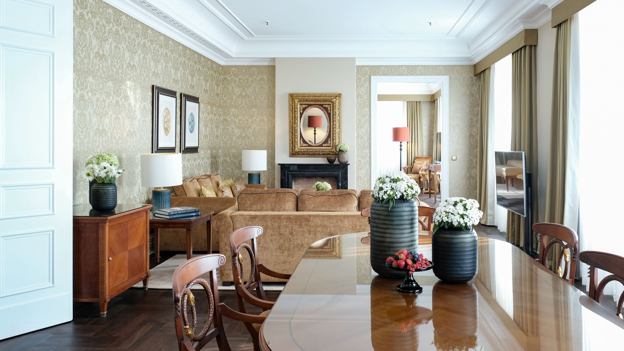 A spacious living room with a large wooden coffee table, a sofa, and a fireplace. There are framed pictures on the wall and a large mirror reflecting the room.