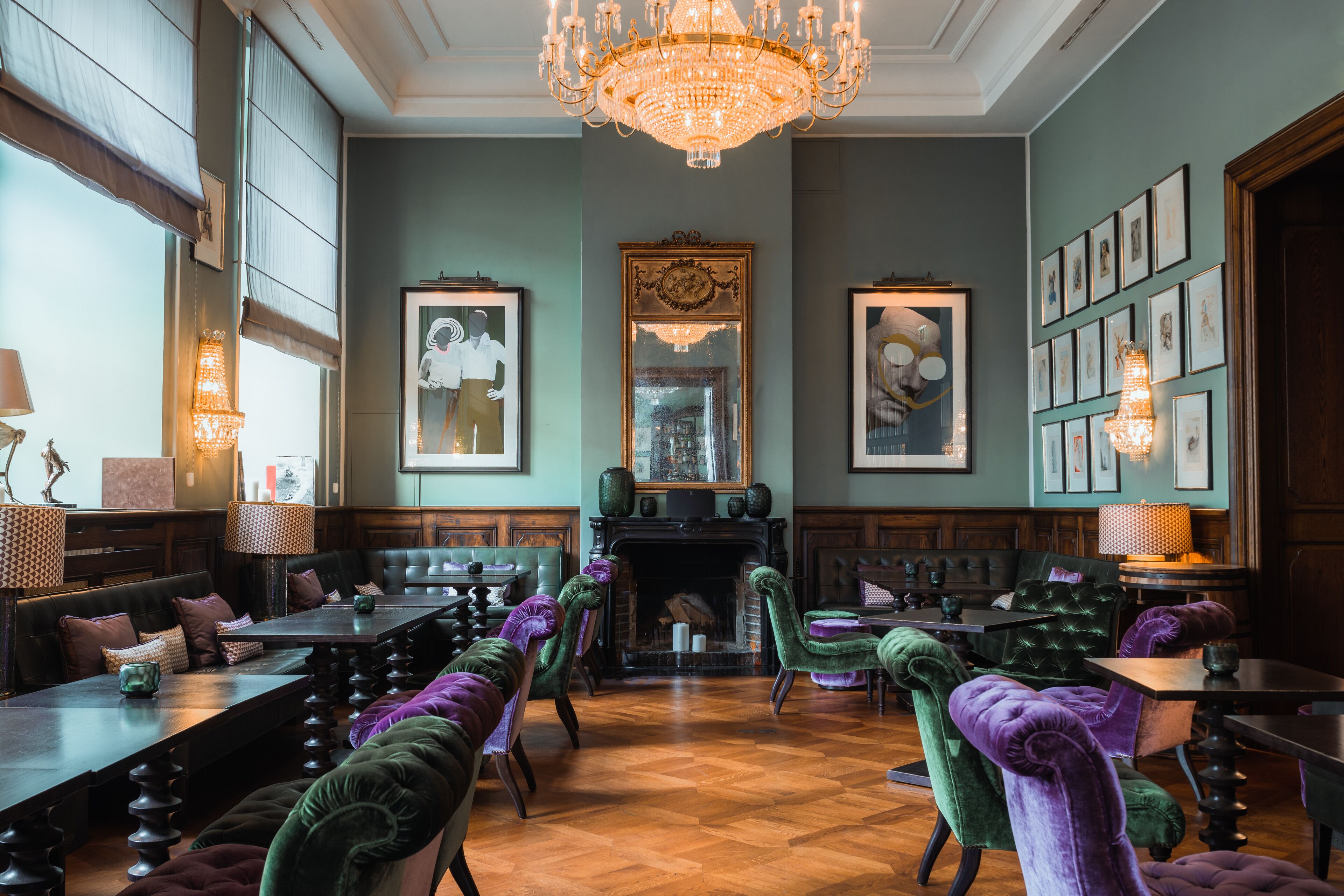 Un comedor amplio con mesas de madera altas y cómodas sillas tapizadas en verde y morado, un gran candelabro y obras de arte enmarcadas en las paredes.