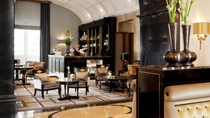 Un hall d’entrée luxueux avec un plafond haut, un sol en marbre et un mobilier élégant. On y trouve un bar avec un barman, et l’espace est bien éclairé grâce à un éclairage d’ambiance.