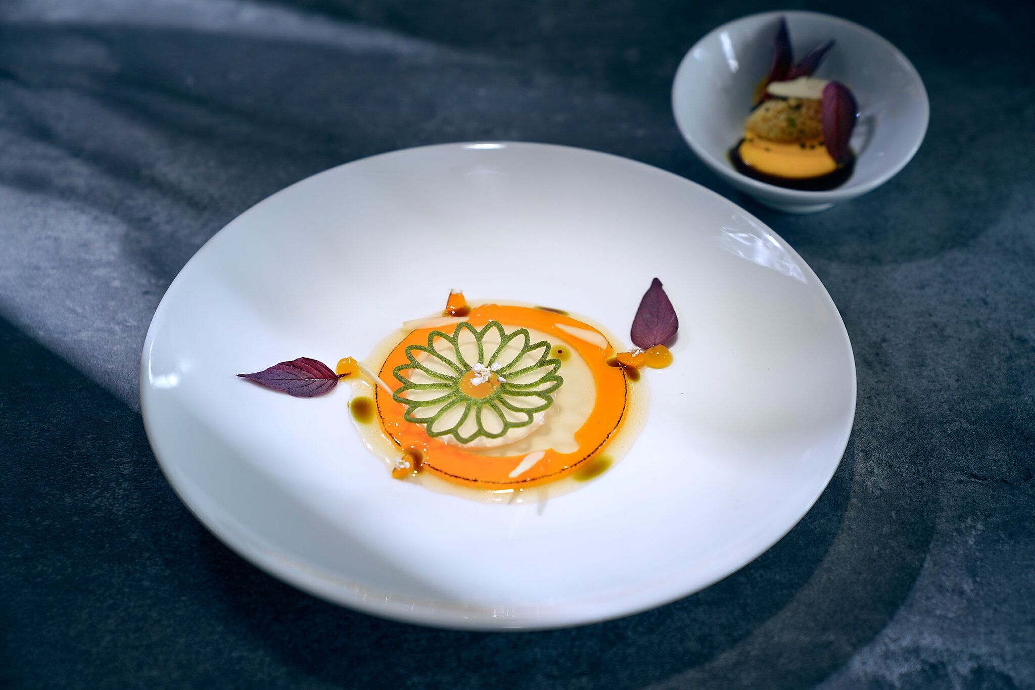 Ein weißer Teller mit einem Dessert mit floralem Muster, dekoriert mit Blättern und Soße, auf einer dunklen Oberfläche.