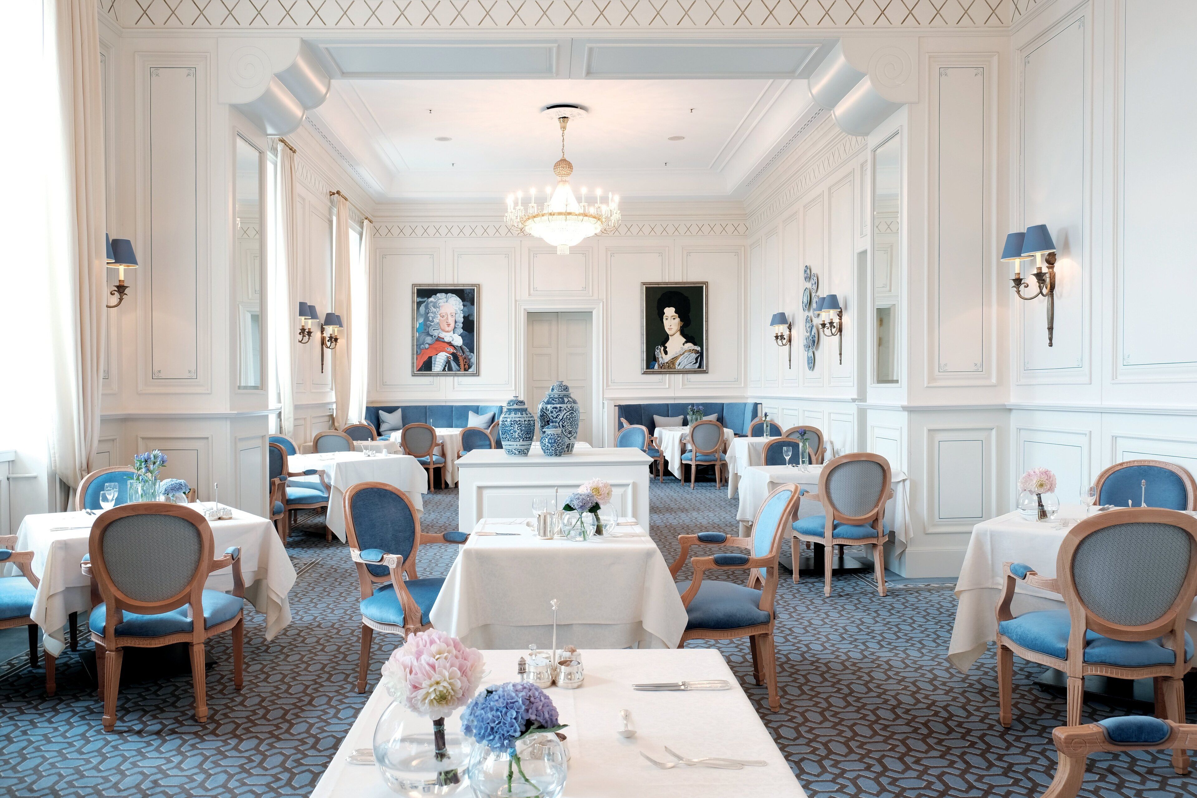 A formal dining room with tables set for service, elegant chairs, and a chandelier.