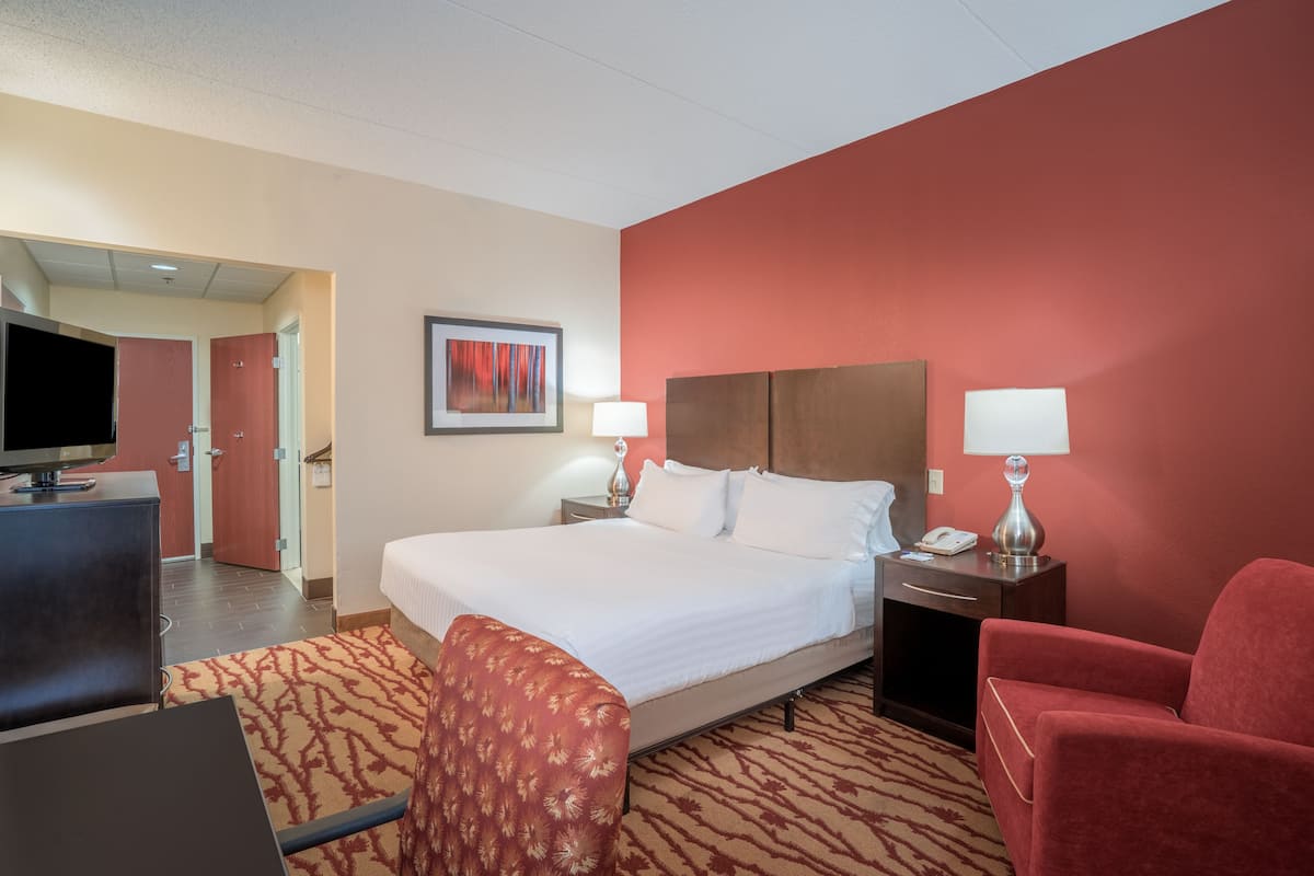 A hotel room with a large bed, two bedside tables with lamps, a desk with a computer, a TV, and a red armchair.