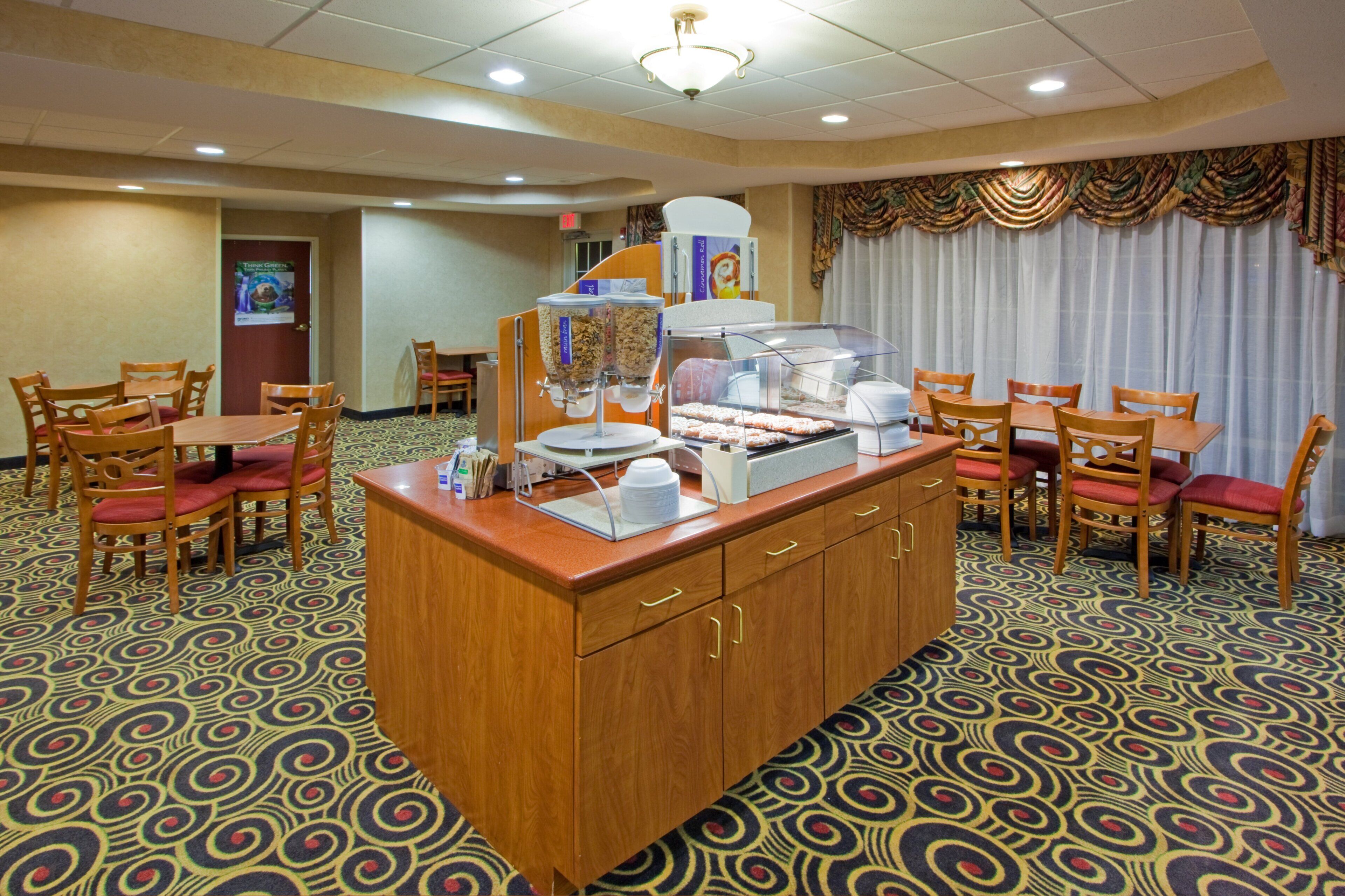 Espaço de café da manhã de hotel, com balcão de buffet, cadeiras e carpete estampado.