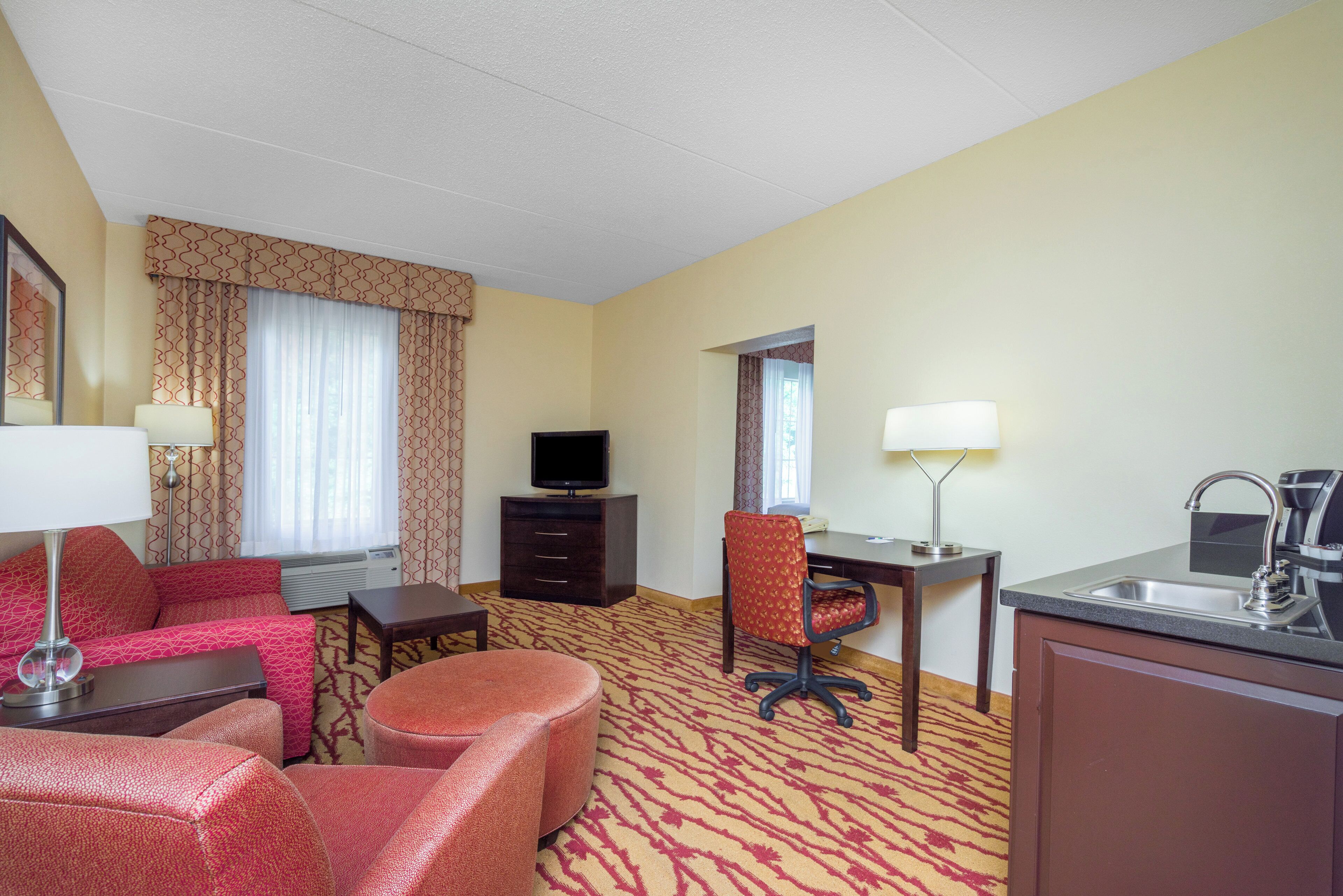 A hotel room with a sofa, a desk, a chair, a television, a desk lamp, and a kitchenette.