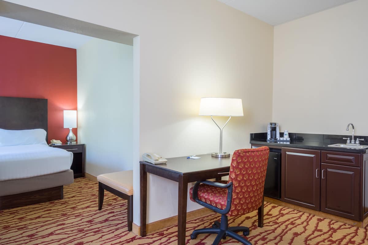 A hotel room with a bed, desk, chair, lamp, and kitchenette.