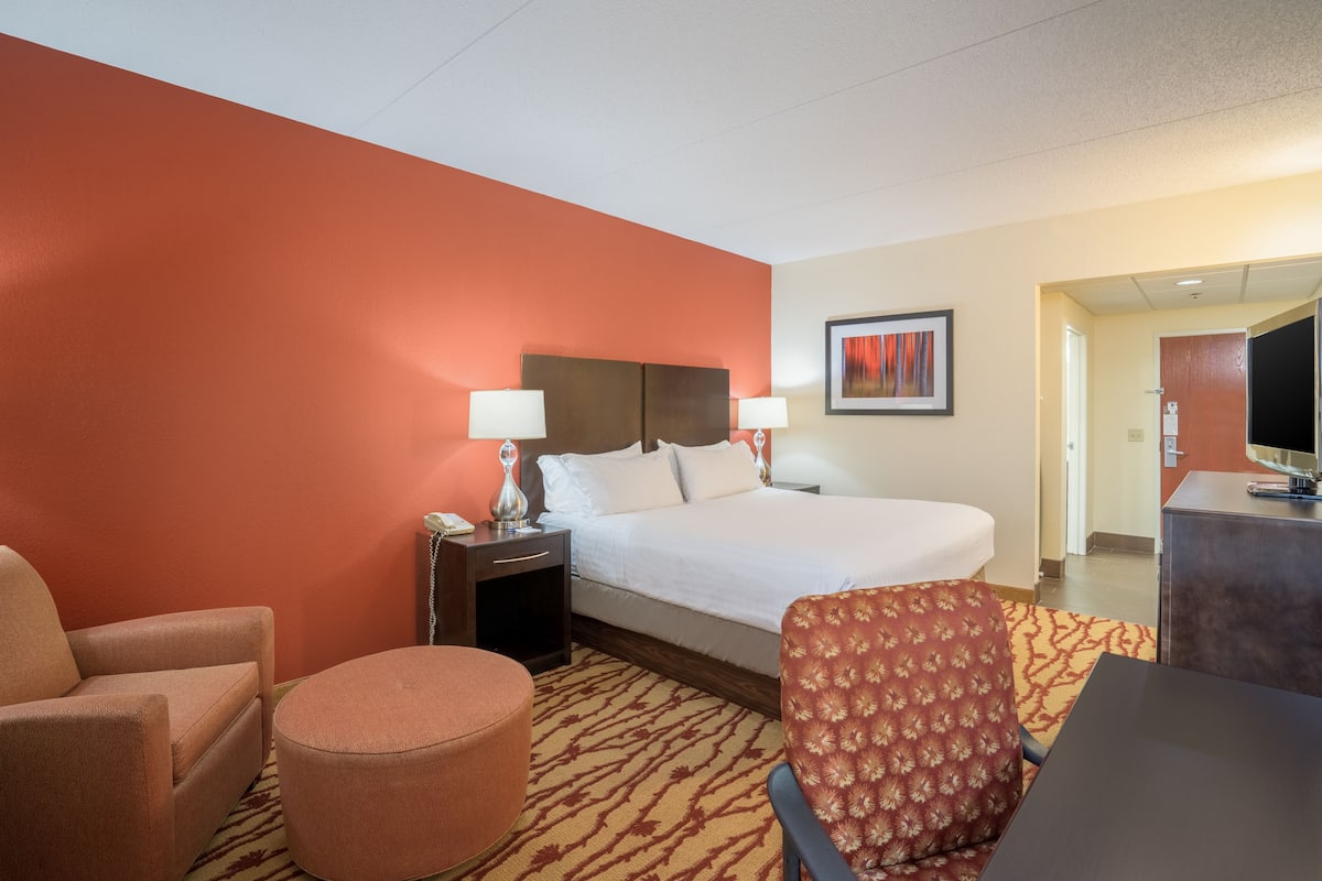 A hotel room with a large bed, two bedside tables with lamps, a chair, a small ottoman, and a desk with a television.