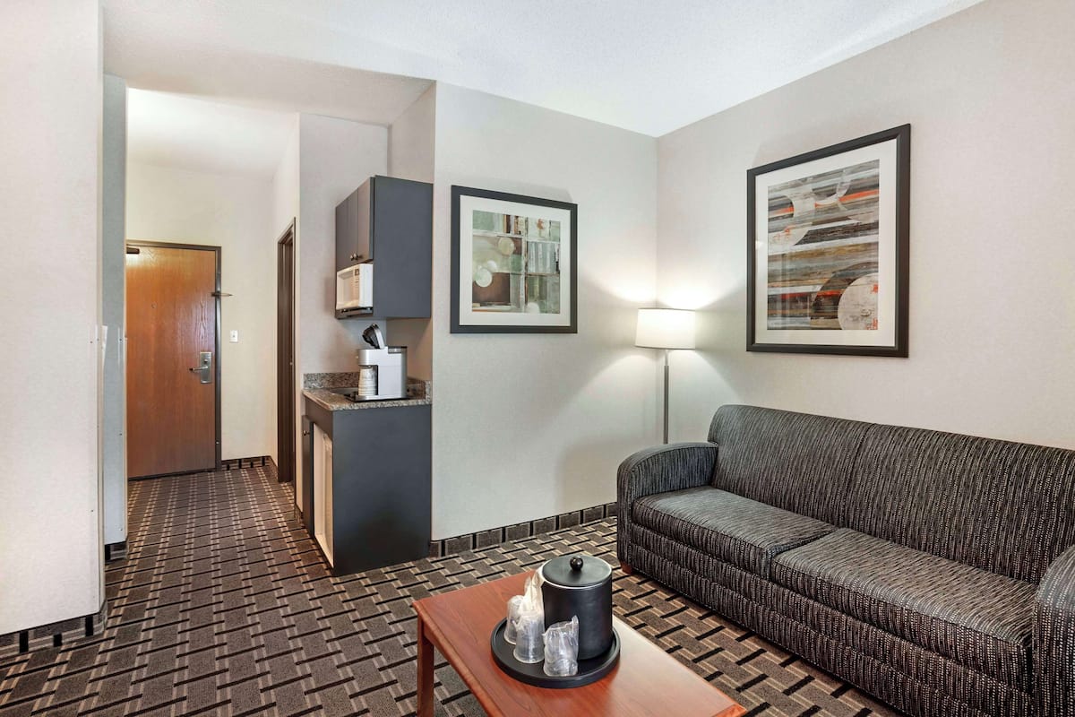 A hotel room with a sofa, a small table with a lamp, a kitchen area, and two framed pictures on the wall.
