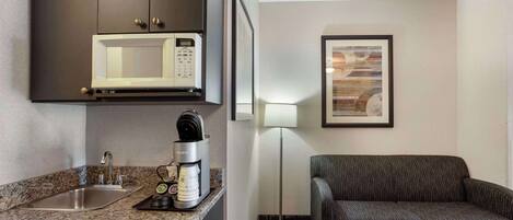 Habitación de hotel compacta con kitchenette, microondas, cafetera, sofá y una lámpara de pie.