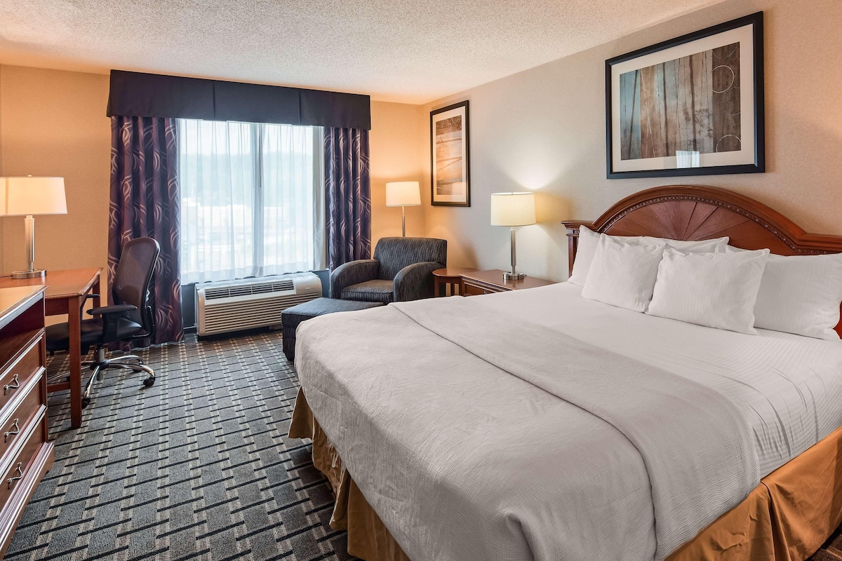 A hotel room with a large bed, a desk with a lamp, a chair, an air conditioning unit, and a framed picture on the wall.