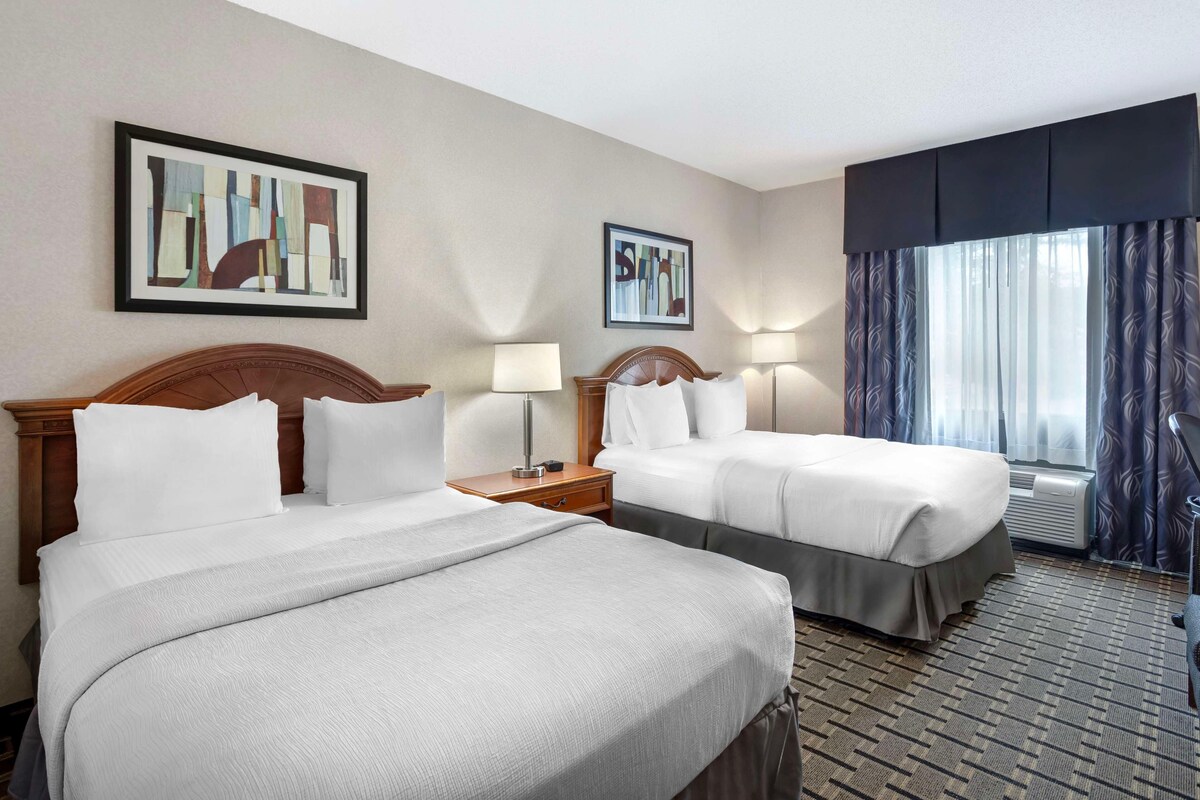 A hotel room with two beds, a nightstand with a lamp, a window with curtains, and two framed artworks on the wall.