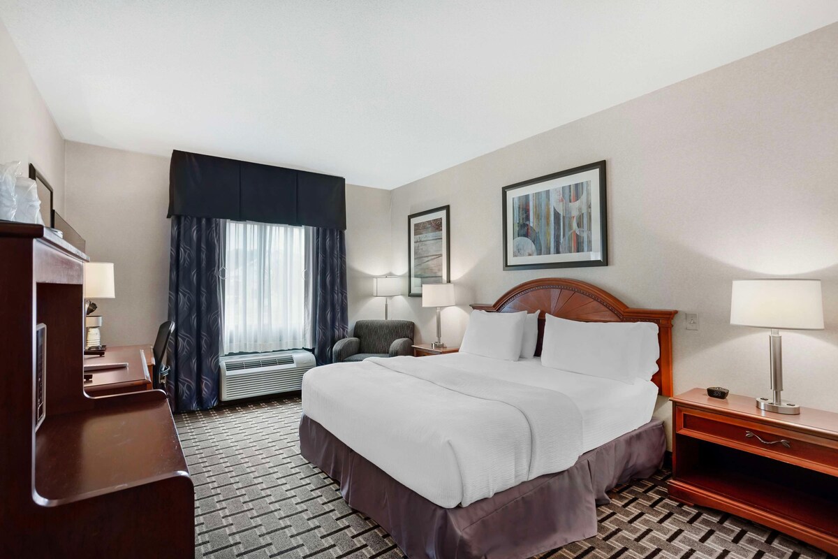 A hotel room with a large bed, a desk, a chair, a nightstand, and a window with curtains.