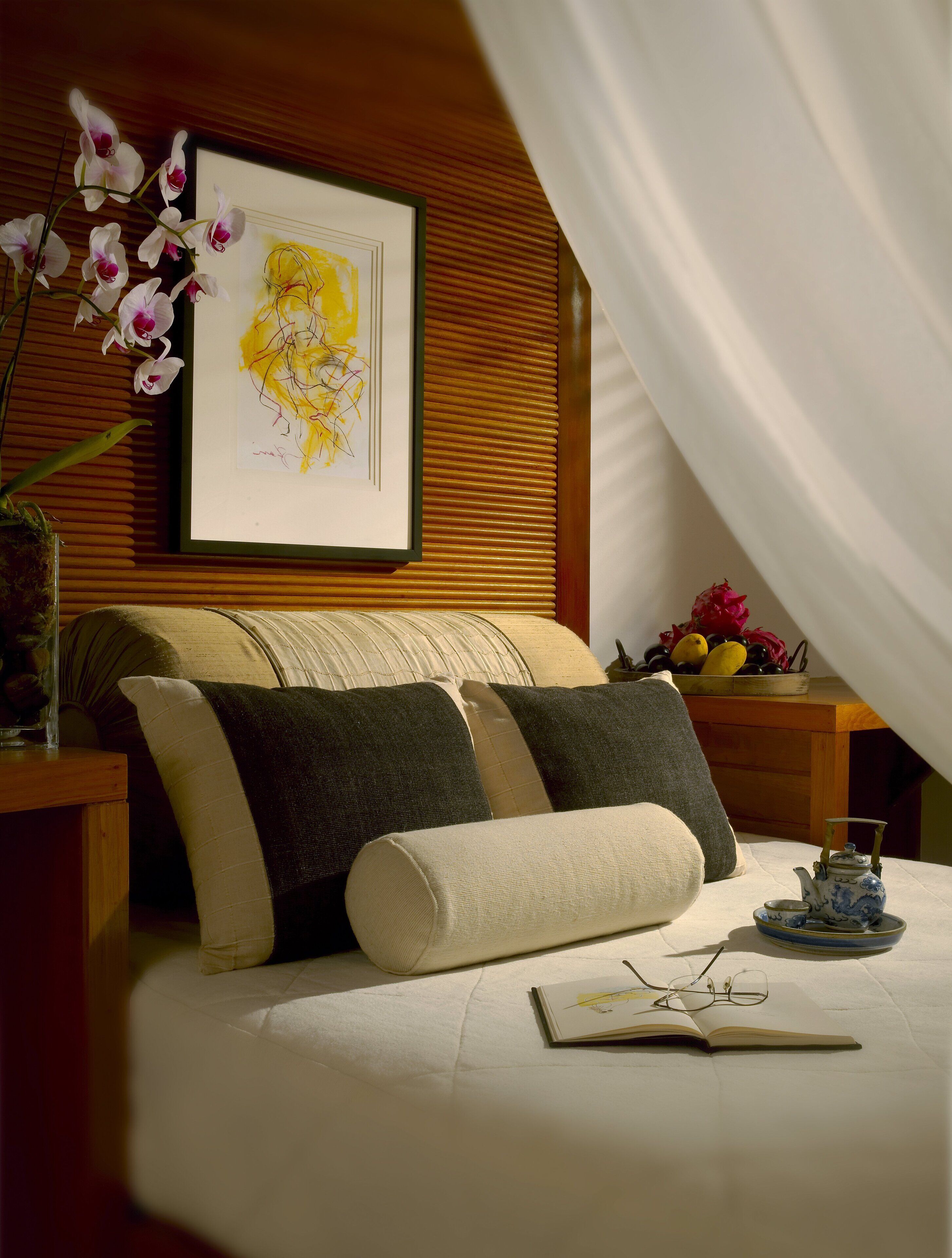 Un dormitorio con una cama, almohadas, una mesita de noche con una tetera, una obra de arte enmarco y un jarrón con flores.