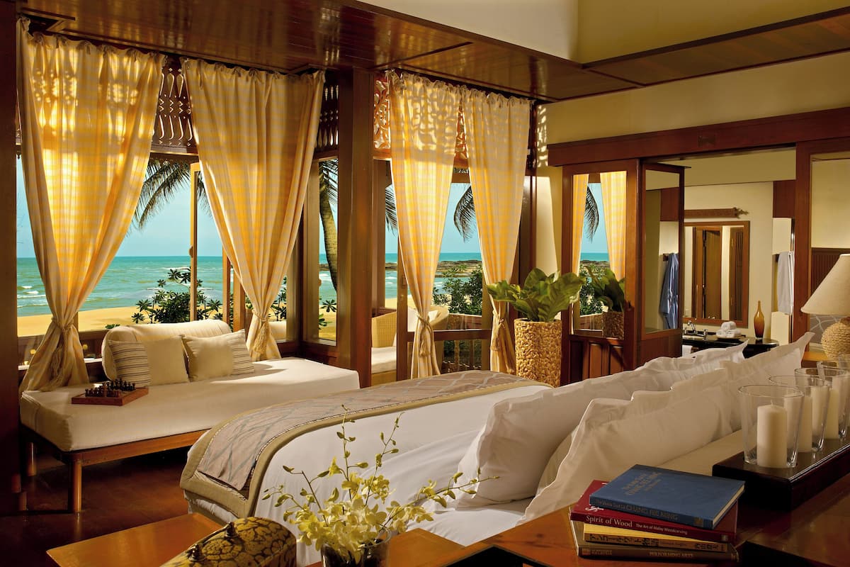 A bedroom with a large bed, a bedside table with books and a vase of flowers, and a view of the beach.