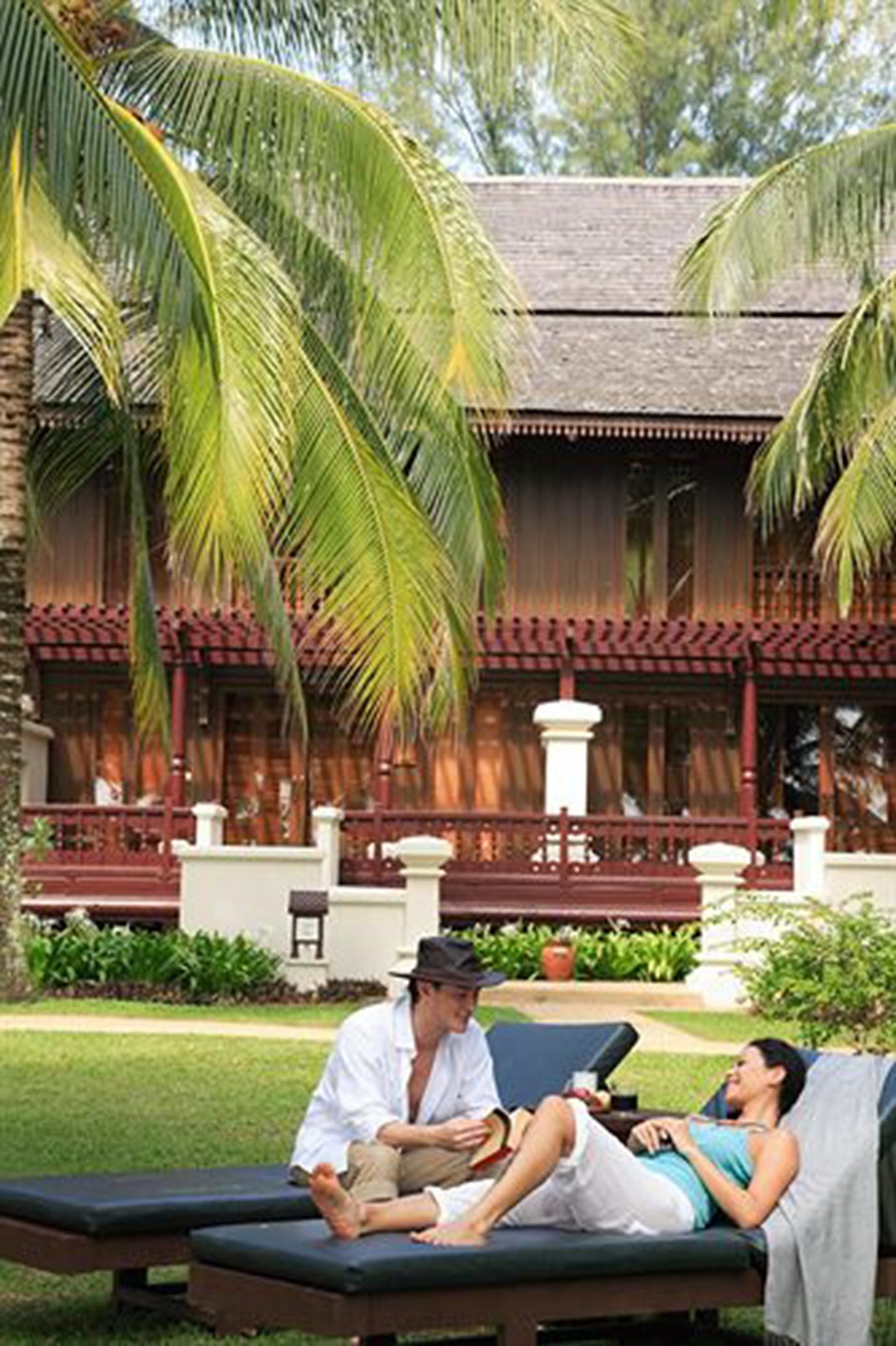 A traditional wooden building with a balcony, palm trees, and two lounge chairs with people relaxing.