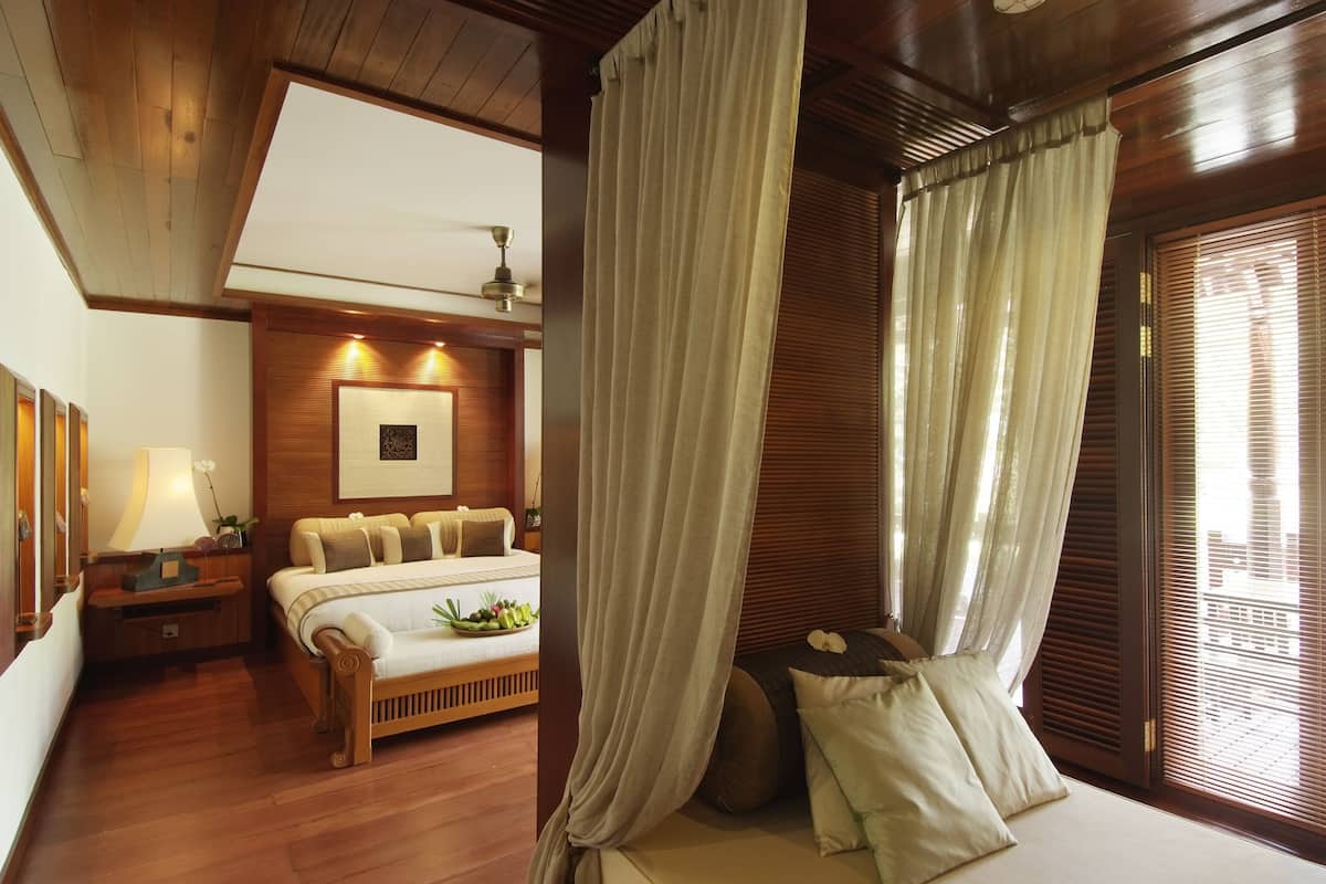 A bedroom with a wooden ceiling, a bed with white bedding, a bedside table with a lamp, and a window with blinds.