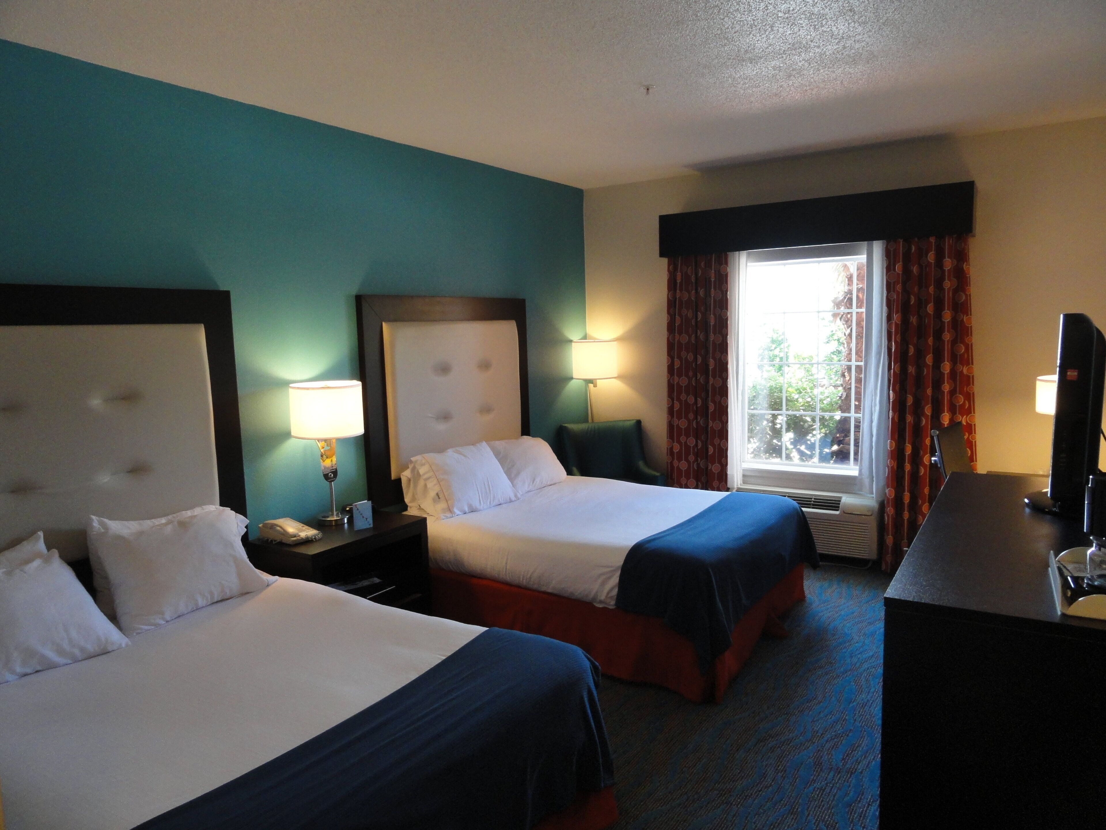A hotel room with two beds, a desk, a television, and a window with curtains.
