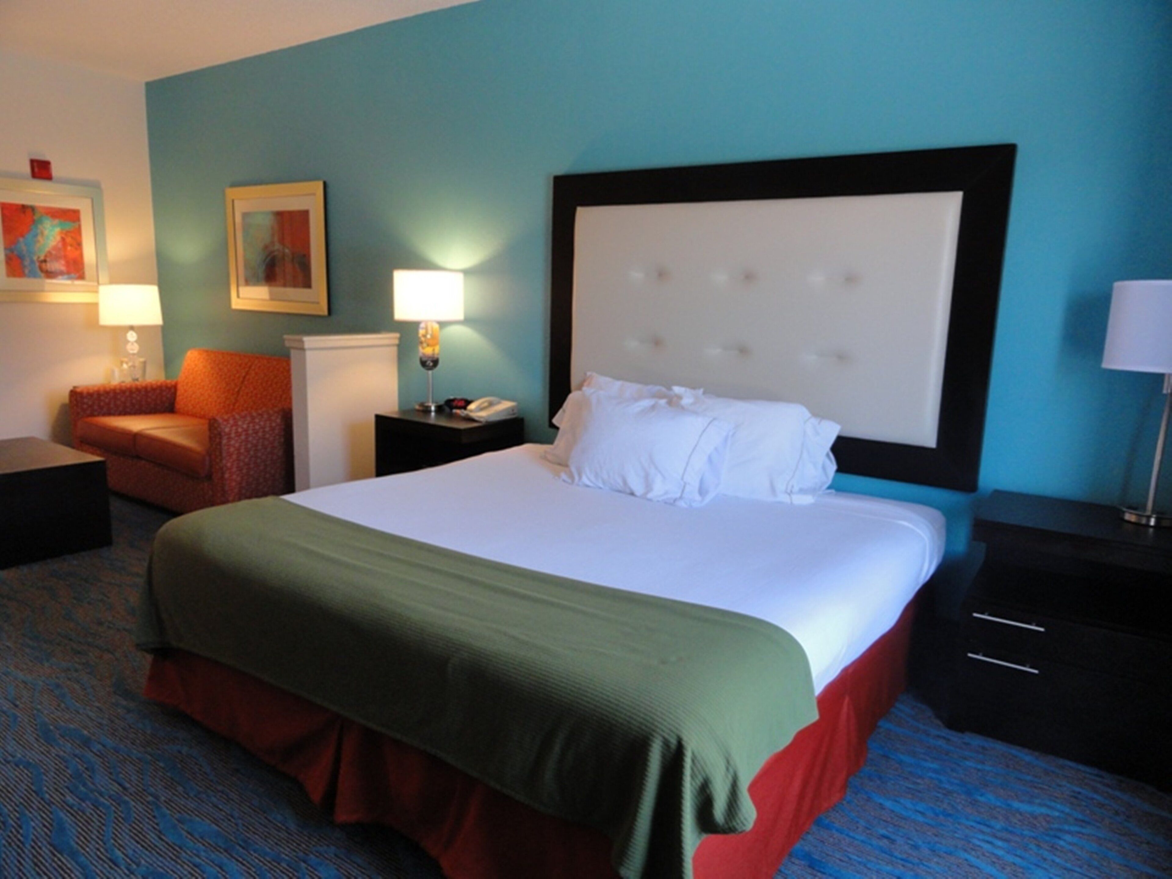 Habitación de hotel con una cama grande, una mesita de noche, una lámpara, una silla y un cuadro en la pared.