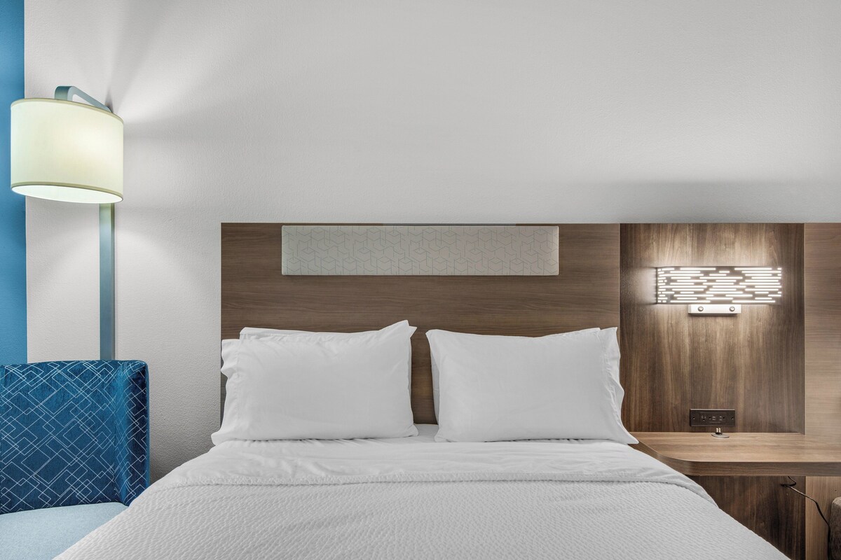 Een hotelkamer met een bed, nachtkastje, lamp en een bedieningspaneel aan de muur.