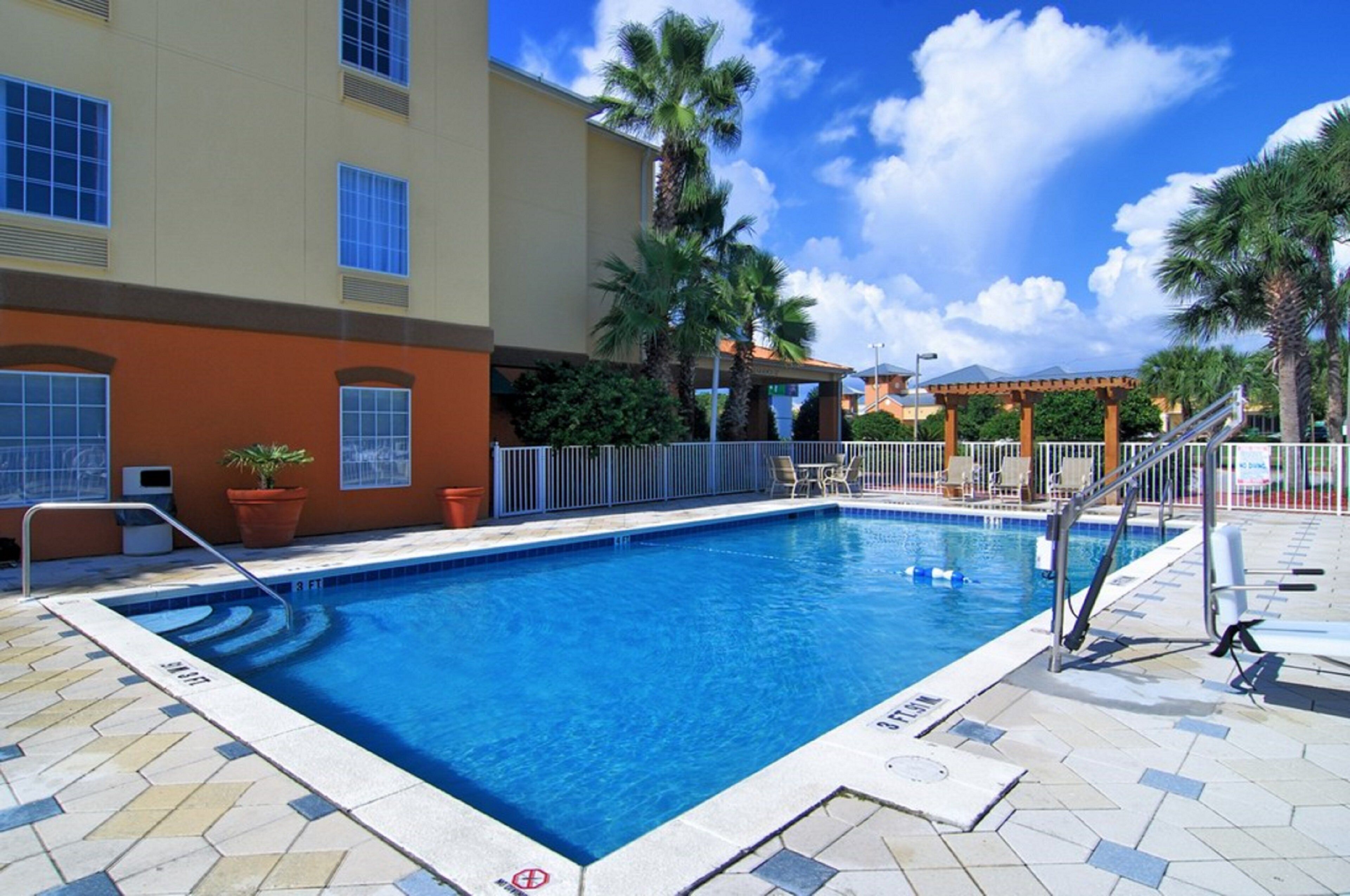 Une piscine dont la profondeur maximale est indiquée à 3 pieds, entourée d’une terrasse carrelée, de palmiers et d’un bâtiment doté de nombreuses fenêtres.