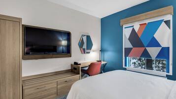 A hotel room with a bed, a desk, a television, a mirror, and a window with a geometric pattern.