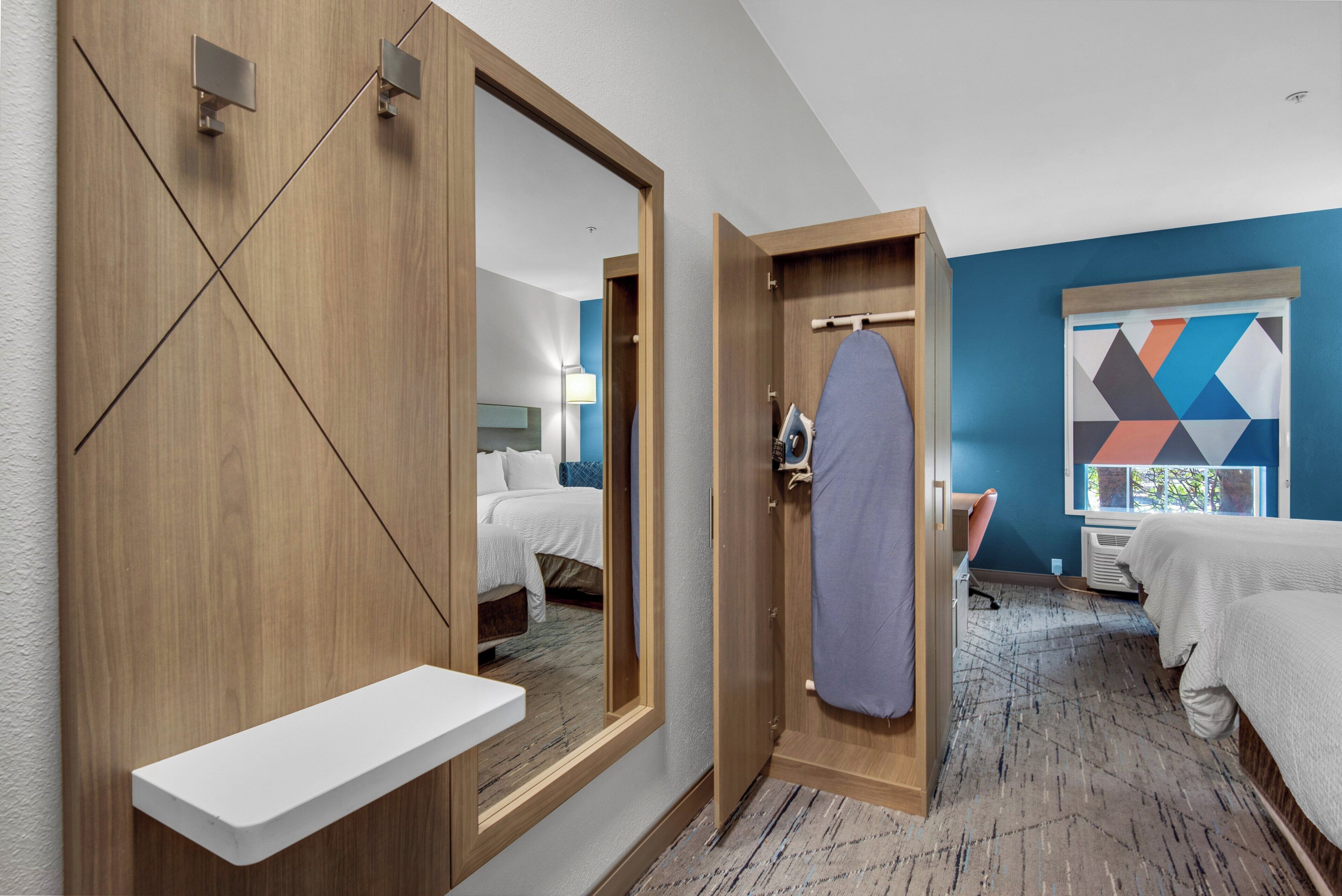 A hotel room with a large mirror, a wooden wardrobe, a white bedside table, a bed with white bedding, a blue wall, and a geometric-patterned artwork.
