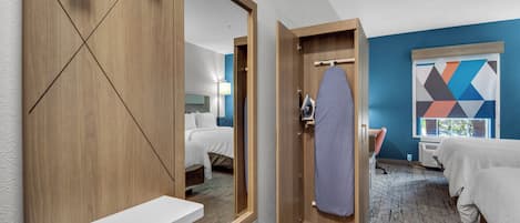 A hotel room with a large mirror, a wooden wardrobe, a white bedside table, a bed with white bedding, a blue wall, and a geometric-patterned artwork.