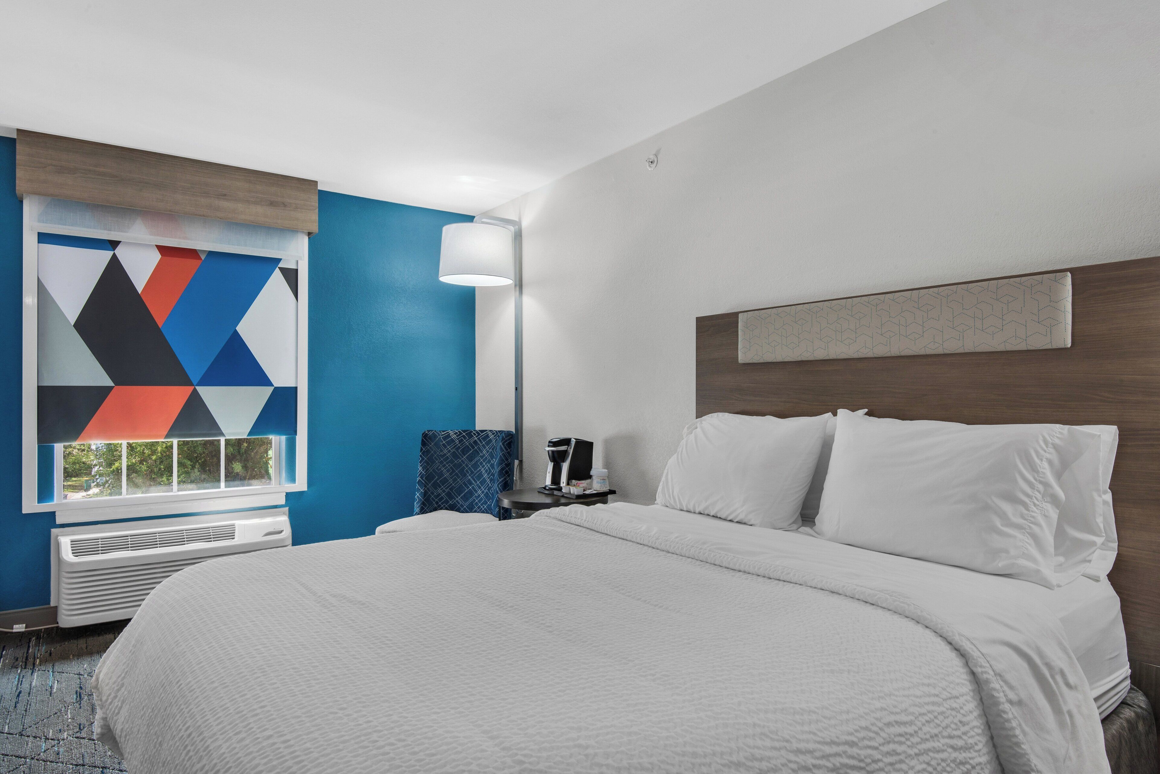 Quarto de hotel com uma cama grande, uma mesinha de cabeceira, um abajur e uma janela com uma obra de arte geométrica na parede.