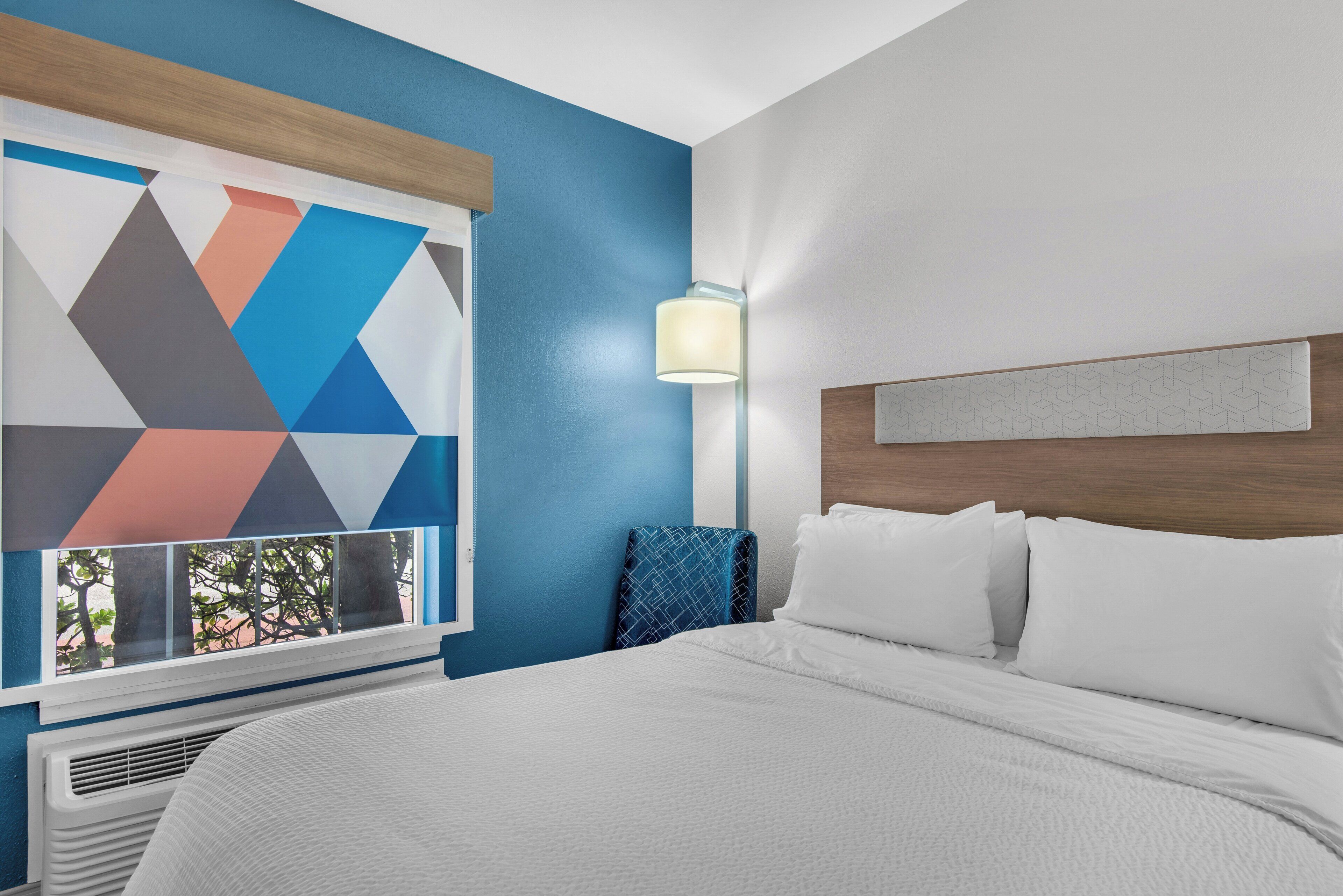Una camera d'albergo con un letto, una parete decorata con arte geometrica, una finestra con persiane e una lampada a parete.