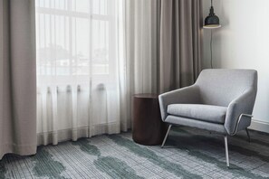 A room with a grey armchair, a round wooden side table, a floor lamp, and a window with sheer curtains. - Hilton Garden Inn Annapolis Downtown (Annapolis)
