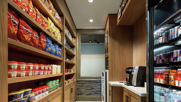A narrow corridor with shelves stocked with snacks and beverages, a refrigerator with drinks, and a coffee machine.