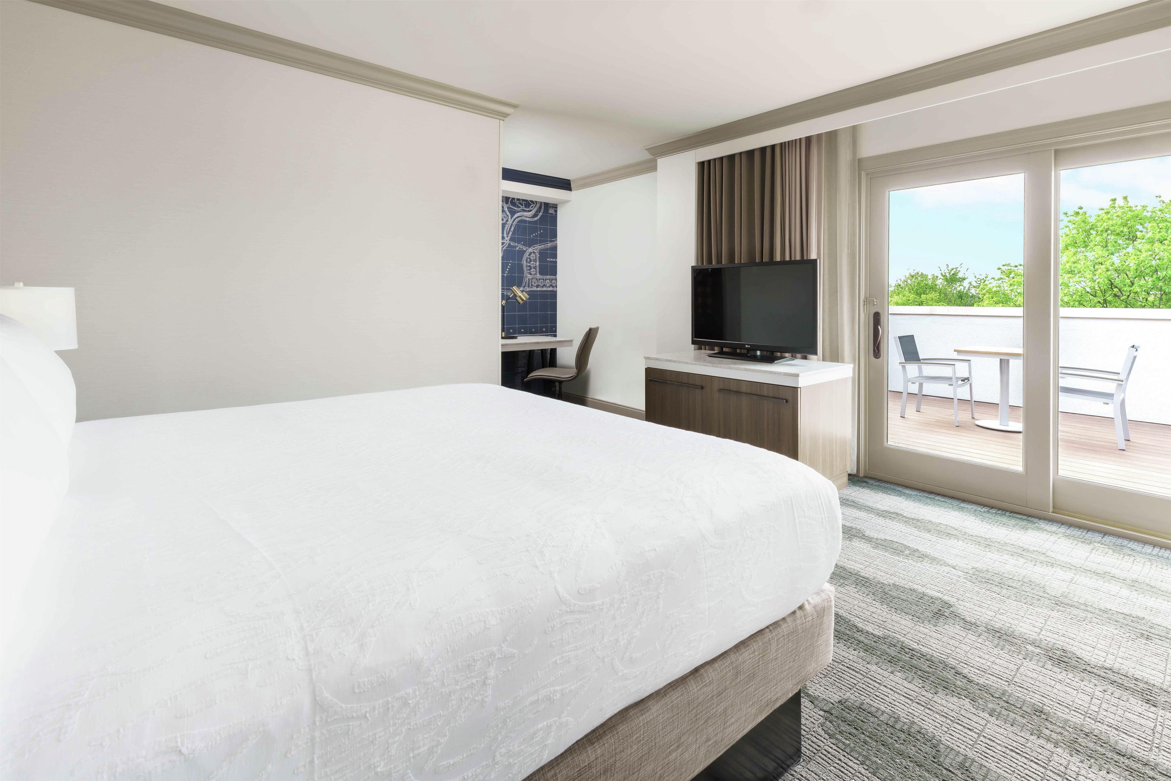 A hotel room with a large bed, a desk with a flat-screen TV, a chair, and a view of the outdoors through a sliding glass door.