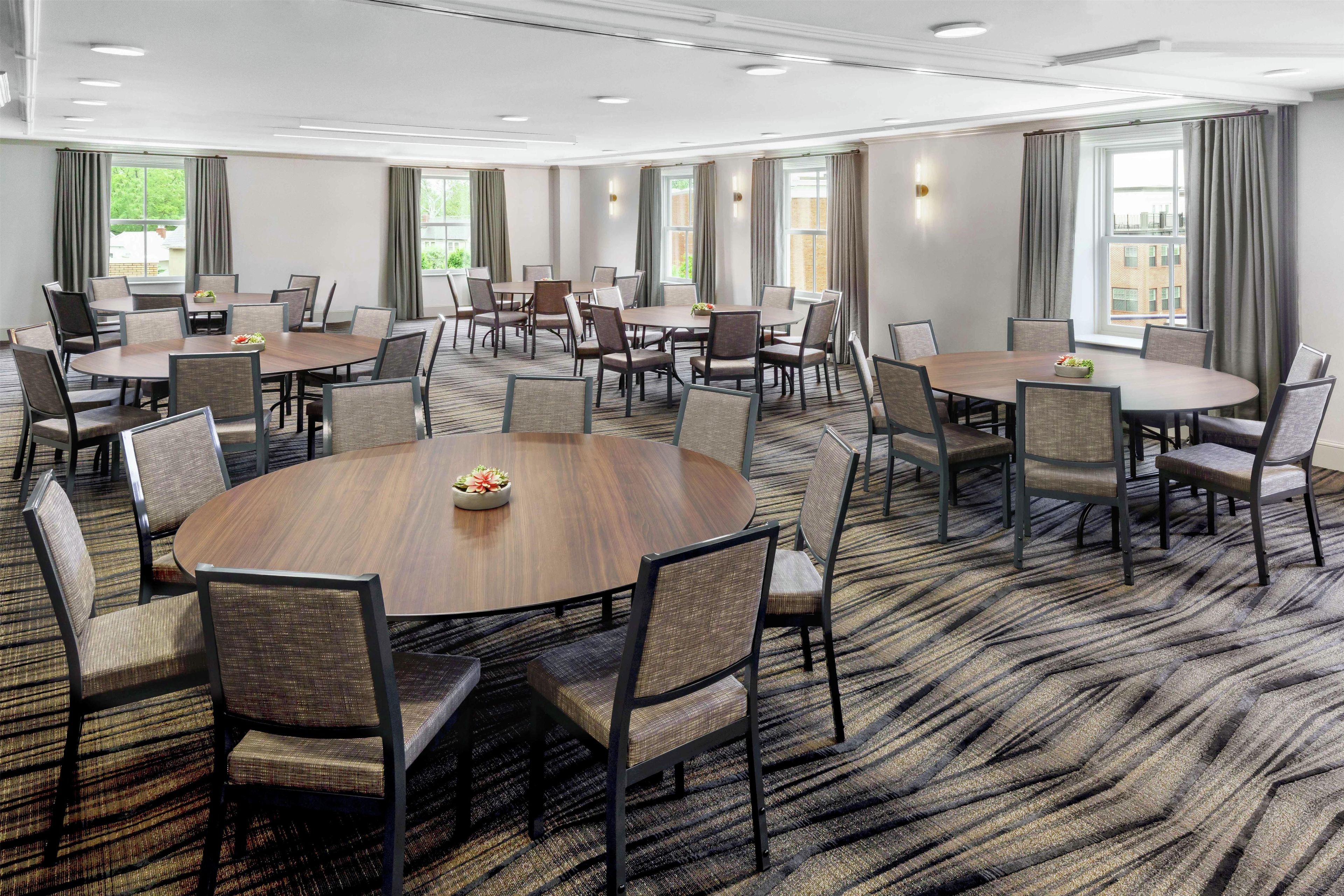 A conference room with multiple round tables and chairs, large windows with curtains, and a patterned carpet.