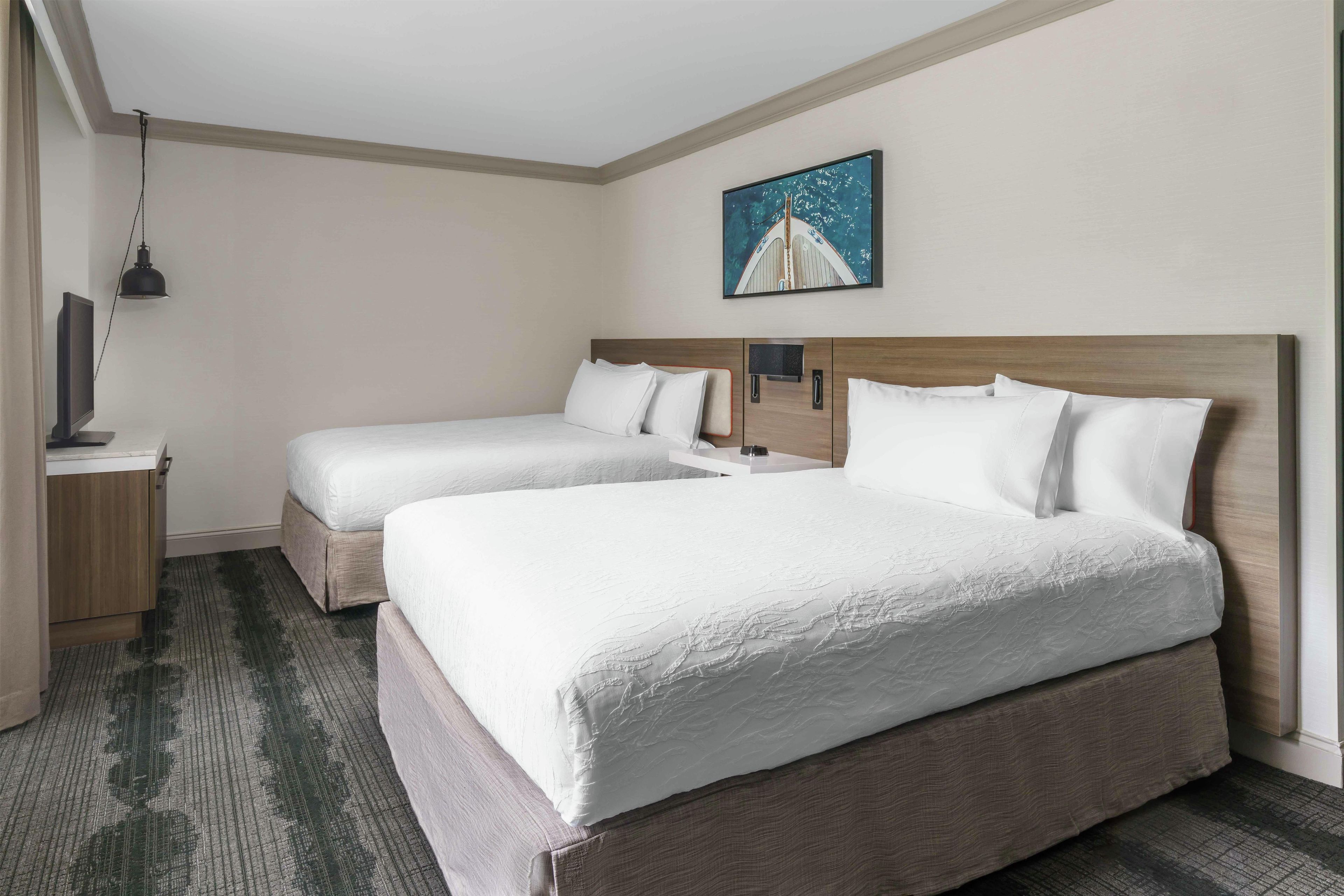 A hotel room with two beds, a flat-screen TV, a painting, and a desk with a lamp.