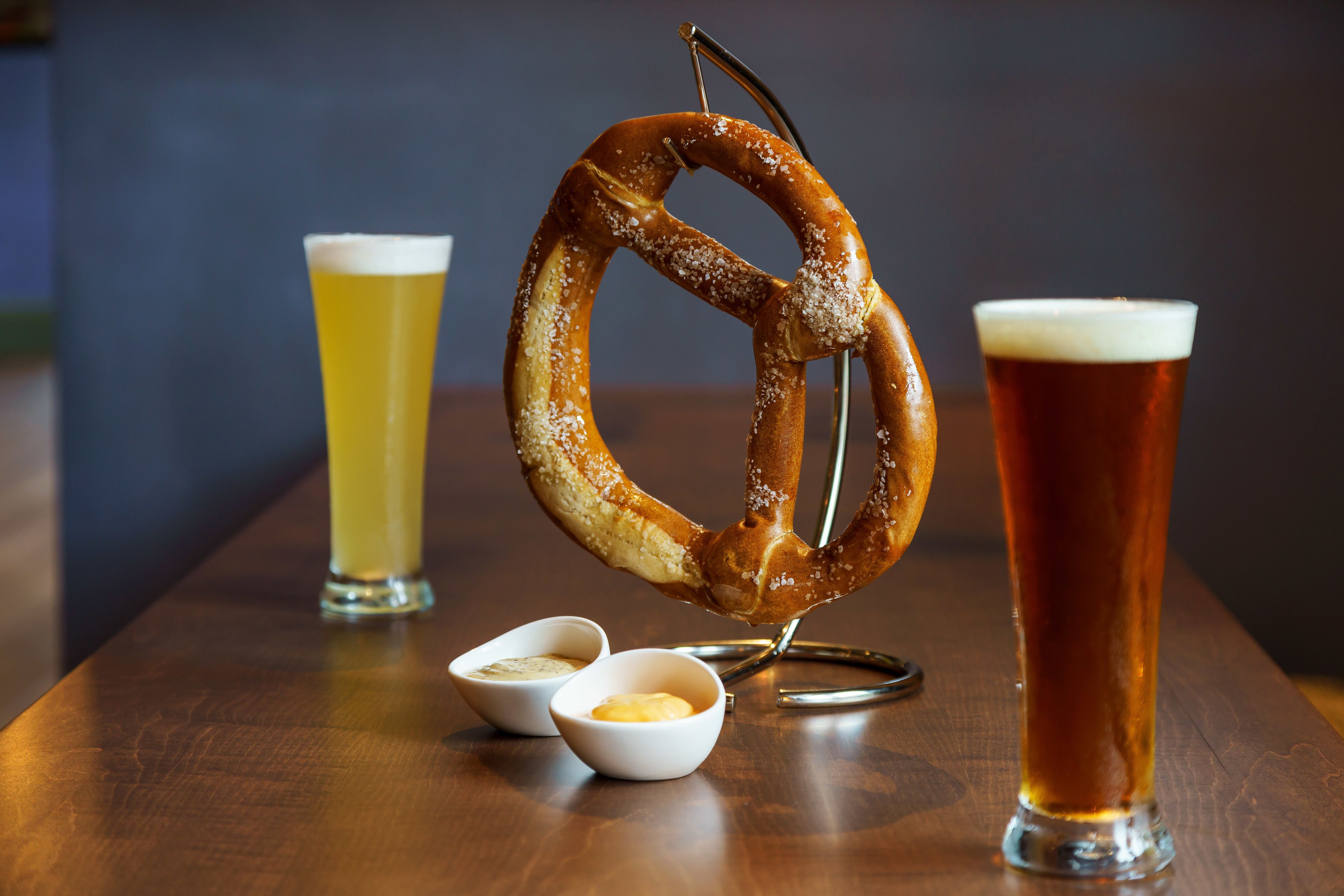 A pretzel on a hook, two small bowls of butter, and two glasses of beer on a wooden table.