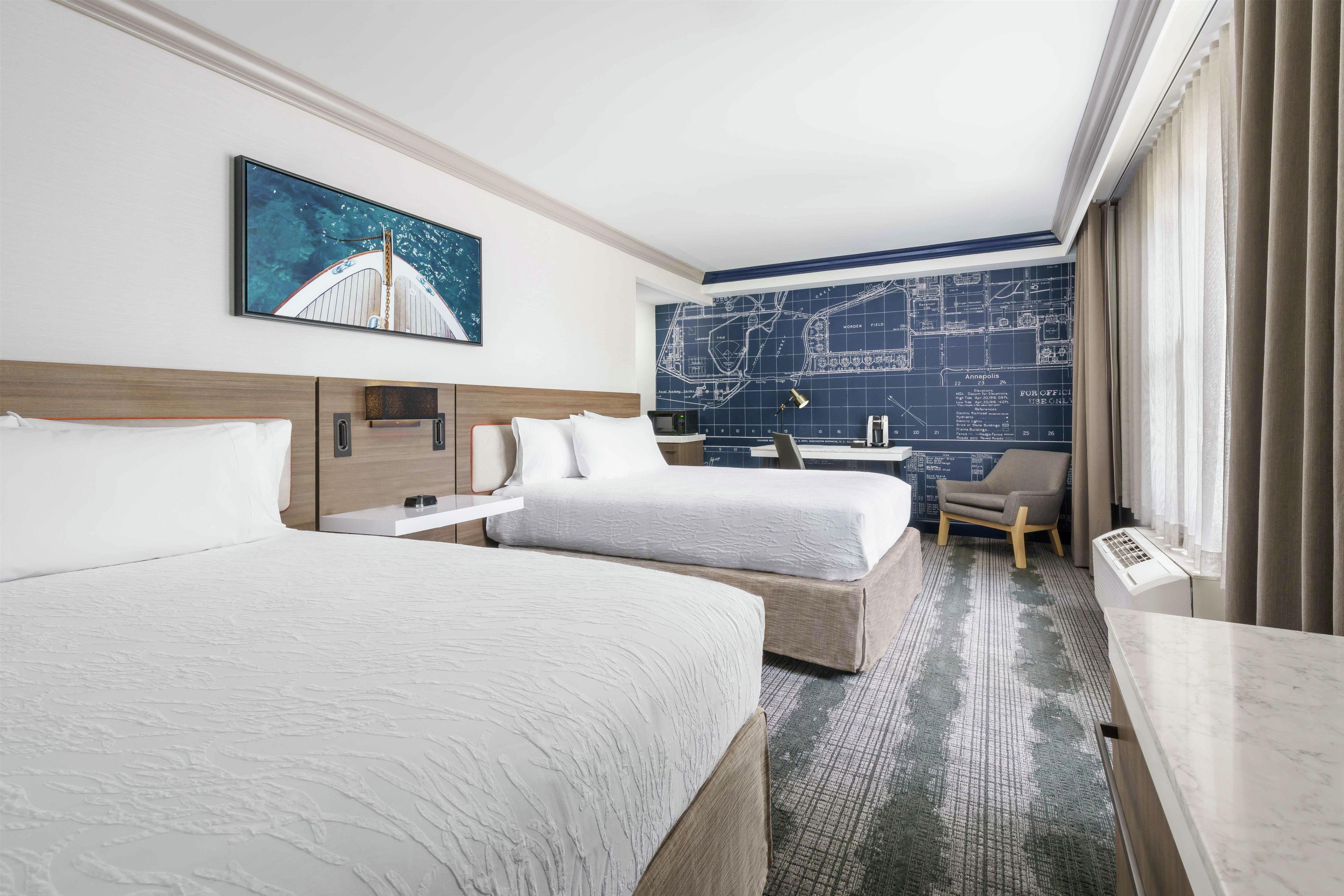 Habitación de hotel con dos camas, un escritorio, una silla, un mapa en la pared y un ventanal con cortinas.