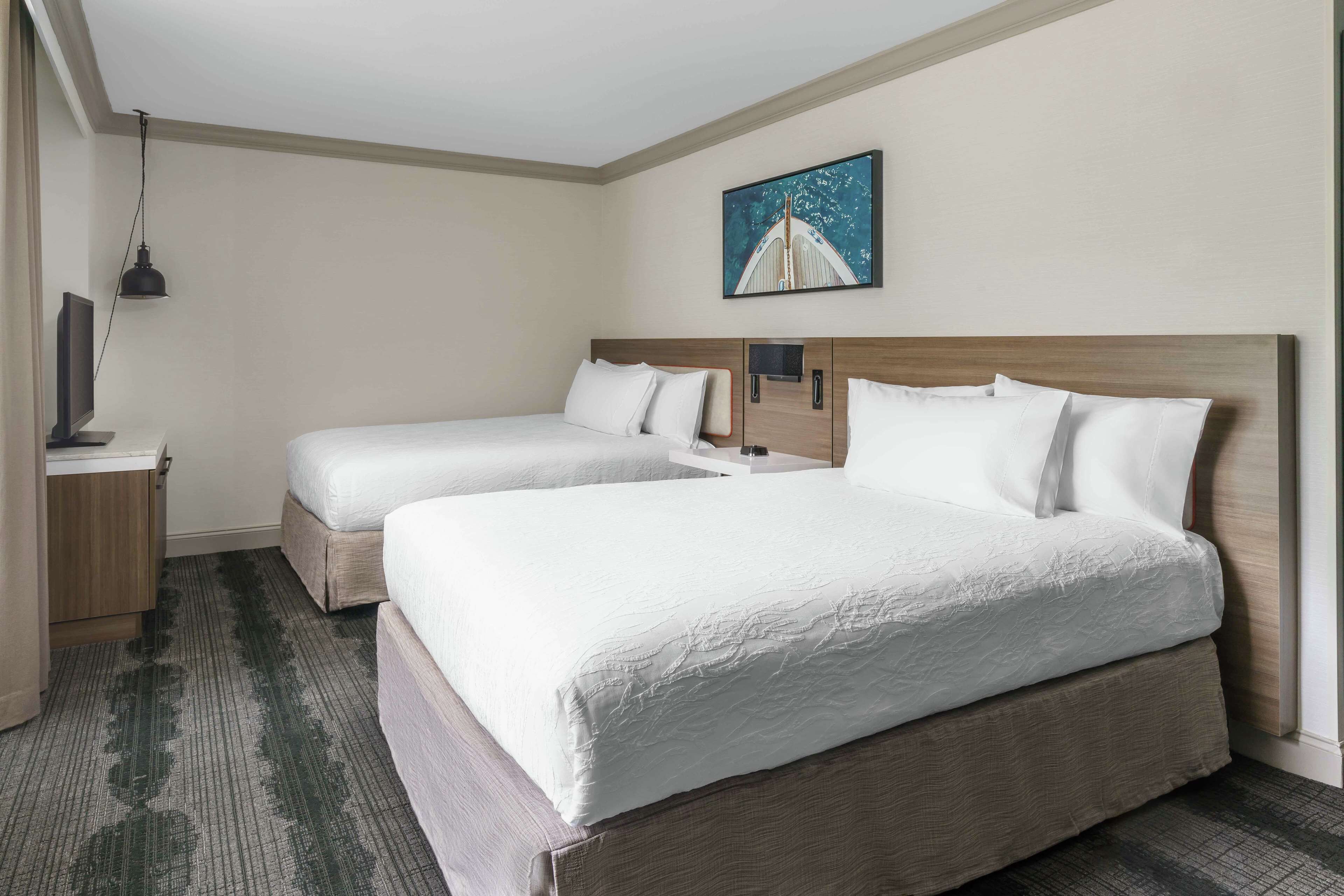 A hotel room with two beds, a flat-screen TV on a wooden nightstand, a painting on the wall, and a carpeted floor.