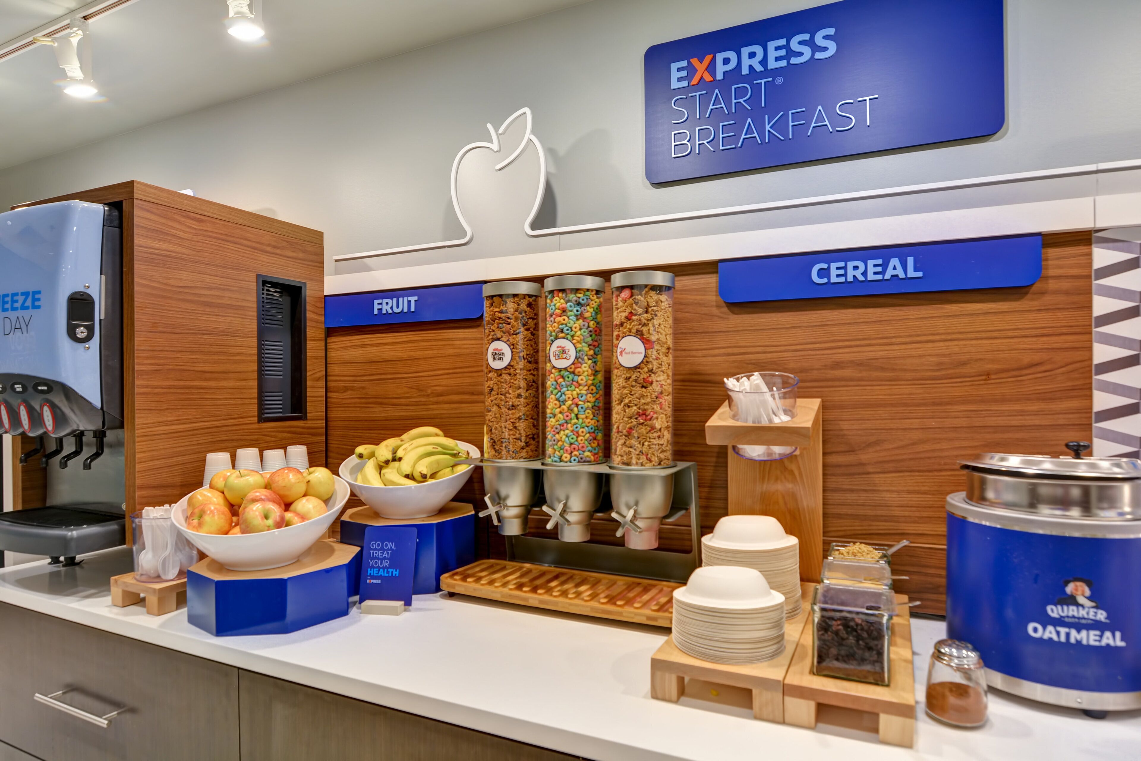A breakfast buffet area with labeled sections for fruit and cereal, including bowls of apples and bananas, and dispensers for Quaker Oatmeal and other cereals.
