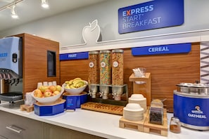 A breakfast buffet area with labeled sections for fruit and cereal, including bowls of apples and bananas, and dispensers for Quaker Oatmeal and other cereals.