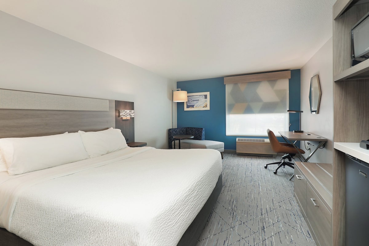 A hotel room with a large bed, a desk, a chair, and a window with blinds.