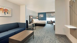 A modern hotel room with a blue sofa, a wooden coffee table, a bed with a headboard, a desk with a chair, and a wall-mounted shelf. - Holiday Inn Express & Suites New Berlin by IHG (New Berlin)