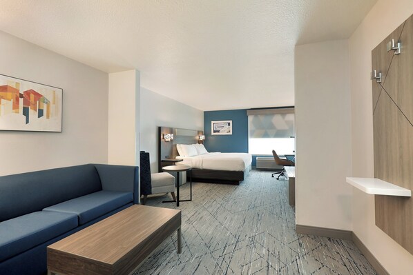 A modern hotel room with a blue sofa, a wooden coffee table, a bed with a headboard, a desk with a chair, and a wall-mounted shelf. - Holiday Inn Express & Suites New Berlin by IHG (New Berlin)