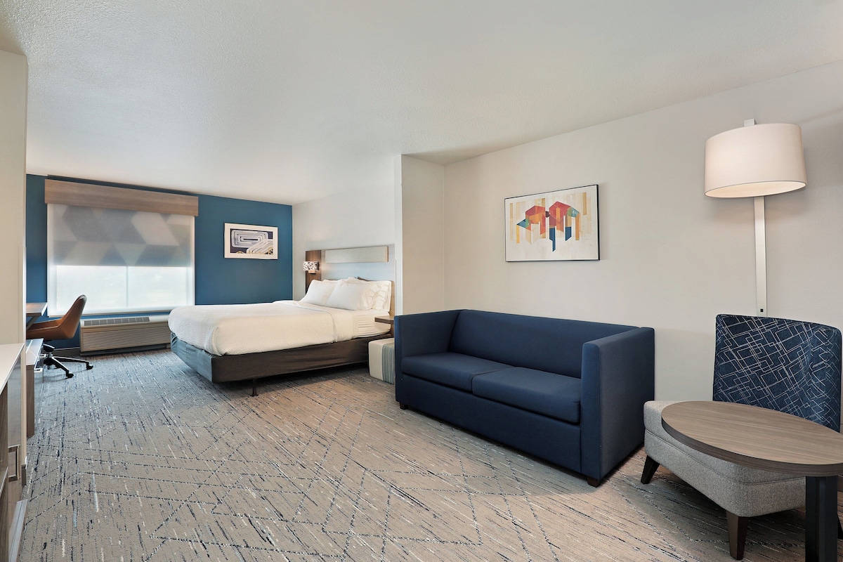 A hotel room with a bed, a blue sofa, a small round table, and a chair. There is a window with blinds, a wall-mounted lamp, and a painting on the wall.