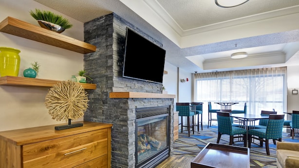 A modern living room with a stone fireplace, wooden shelves, and a flat-screen TV. There is a dining area with blue chairs and a patterned carpet. - Holiday Inn Express & Suites New Berlin by IHG (New Berlin)