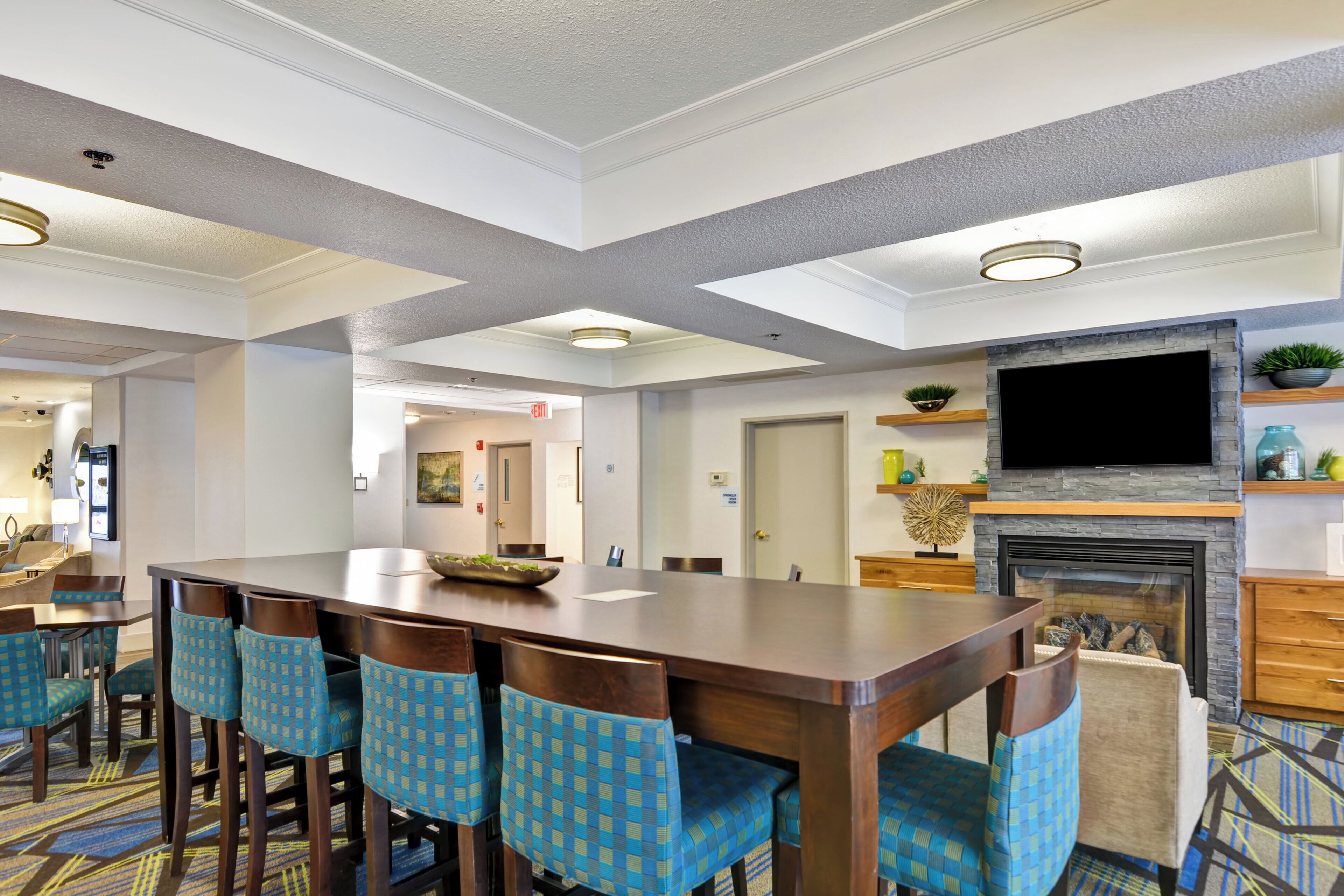 A hotel lobby with a large wooden bar, blue patterned chairs, a flat-screen TV above a fireplace, and a carpeted floor.